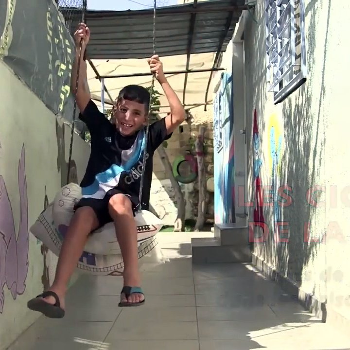 L'image montre un jeune garçon qui se balançe sur une balançoire improvisée. Il semble joyeux et se trouve dans un espace qui semble être un couloir ou une cour. Les murs autour de lui sont décorés avec des peintures murales, dont une représente un chat. Le cadre donne une impression de vie quotidienne dans un endroit où l'art et le jeu se côtoient. On voit également un message en arrière-plan, lié à des thèmes de liberté.