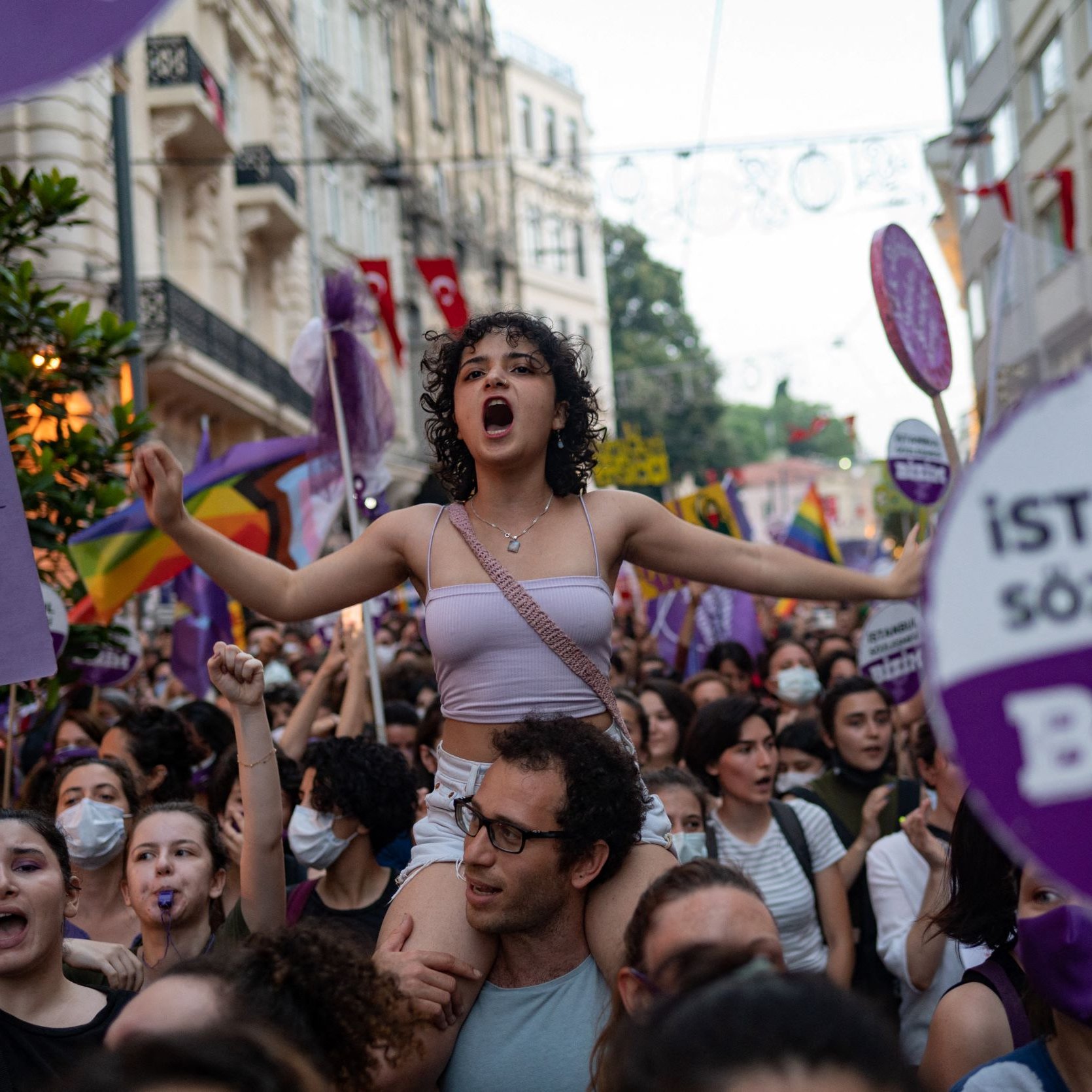 L'immagine mostra un gruppo di persone in una manifestazione per i diritti, con un forte focus sulle tematiche femministe e LGBTQ+. Una giovane donna, in primo piano e con i capelli ricci, è in piedi sulla spalle di un'altra persona e grida, esprimendo la sua passione. Molti partecipanti, prevalentemente donne, sono circondati da bandiere e cartelli colorati, alcuni con slogan che richiamano l'attenzione su temi di uguaglianza e diritti umani. L'atmosfera è vibrante e carica di energia, con un chiaro senso di solidarietà tra i manifestanti.