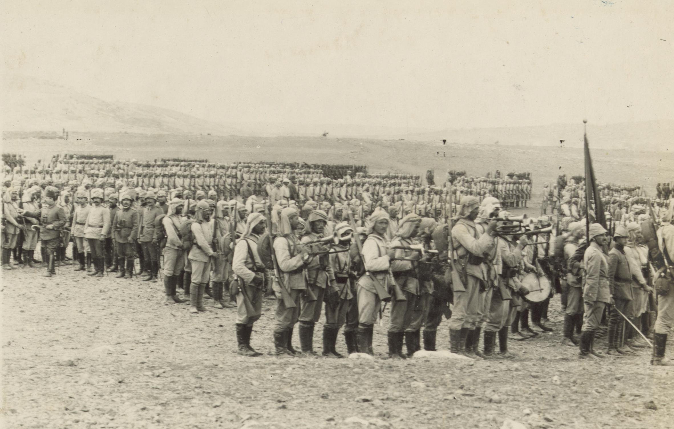 The image depicts a large group of soldiers lined up in a formation during a military event, likely a parade or review. The soldiers are dressed in uniforms typical of early 20th-century military attire, complete with helmets and boots. Some are holding rifles, and there are banners or flags present. In the background, more soldiers can be seen in organized rows, suggesting a significant military presence. The setting appears to be an open field, with hills or mountains visible in the distance, indicating a potentially historic or ceremonial occasion. The image has a monochromatic tone, typical of photographs from that era.