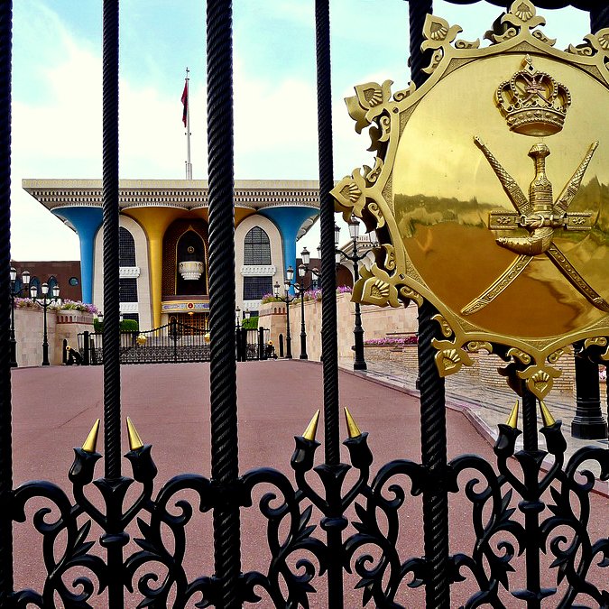 L'image montre un portail d'entrée orné de motifs métalliques élaborés. Au centre, on aperçoit un blason doré avec une couronne et des épées croisées, symbolisant probablement un gouvernement ou une monarchie. À l'arrière-plan, on distingue un bâtiment aux couleurs claires et un drapeau flottant, entouré de pelouses et de fleurs. L'atmosphère dégage une sensation de grandeur et d'autorité.