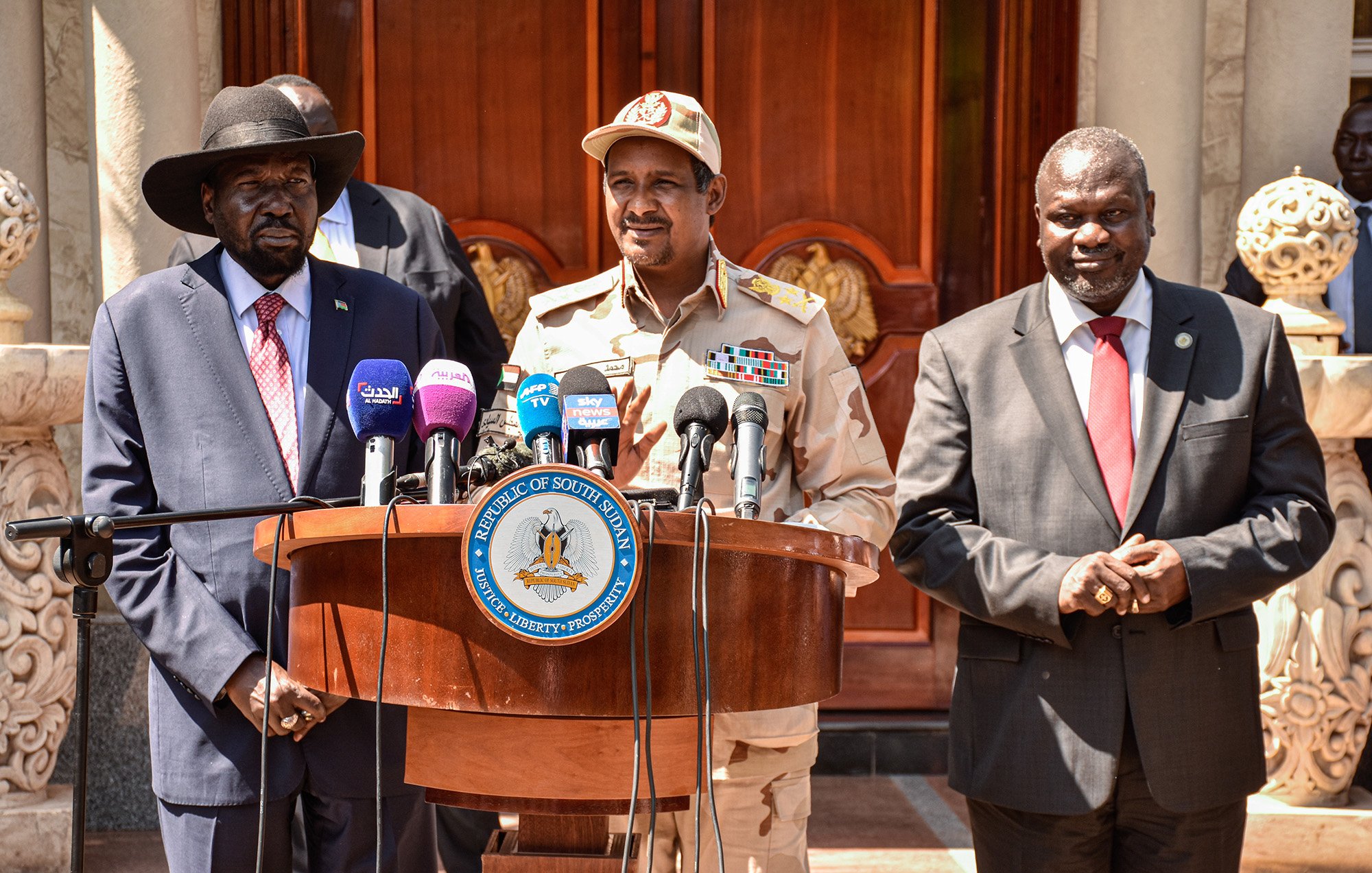 The image shows a group of three men standing behind a wooden podium. The man on the left is wearing a black hat and a suit with a red tie. The man in the center is wearing a military uniform with various insignia and a cap. The man on the right is dressed in a dark suit. They appear to be at an official event, likely addressing the media, with microphones positioned in front of them and a decorative building in the background.