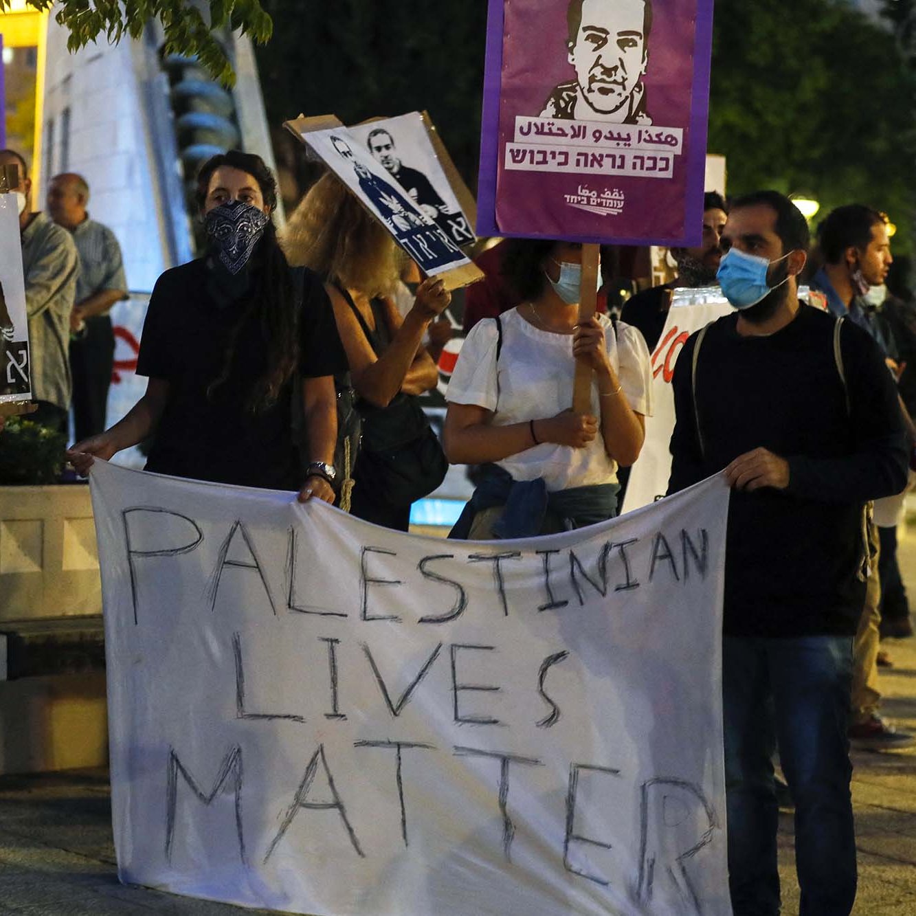در این تصویر، گروهی از مردم در حال برگزاری یک تجمع اعتراضی هستند. آنها تابلوهایی با تصاویر و شعارهایی در دست دارند. یکی از بنرهای بزرگ به زبان انگلیسی می‌گوید "زندگی‌های فلسطینی مهم است". شرکت‌کنندگان به وضوح از ماسک استفاده می‌کنند، و فضای عمومی تجمع نشان‌دهنده یک اعتراض اجتماعی است. در پس‌زمینه، درختان و یک محیط شهری قابل مشاهده است.