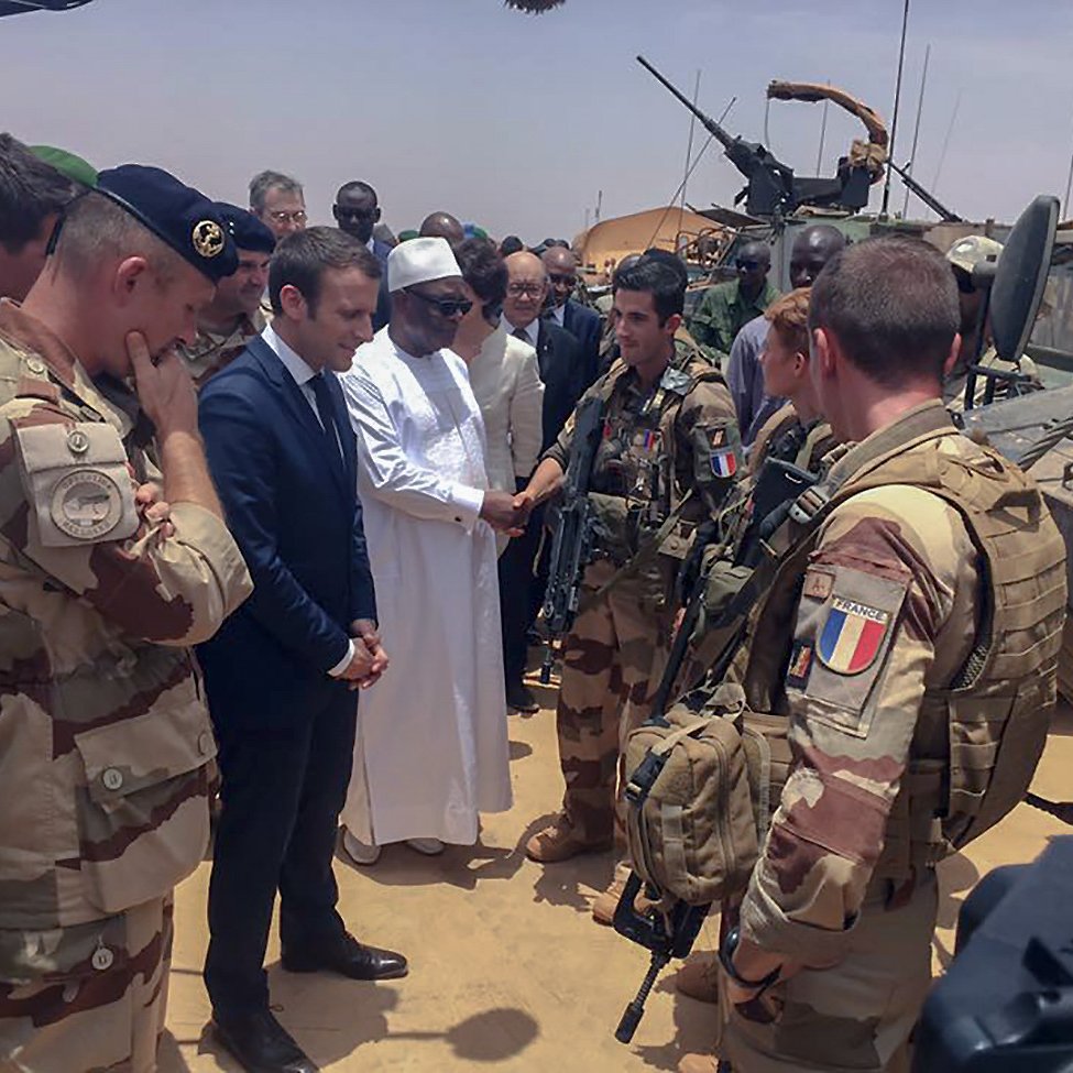 تظهر الصورة مجموعة من الجنود والعسكريين في منطقة صحراوية. في الوسط، يوجد رجل يرتدي لباسًا تقليديًا أبيض، بصحبة شخصية أخرى يبدو أنها رسمية. العسكريون يرتدون زيًا عسكريًا ويحملون الأسلحة، بينما يظهر في الخلفية بعض المركبات العسكرية. الأجواء تبدو جدية، والمشهد يوحي بلقاء رسمي أو زيارة تفقدية.