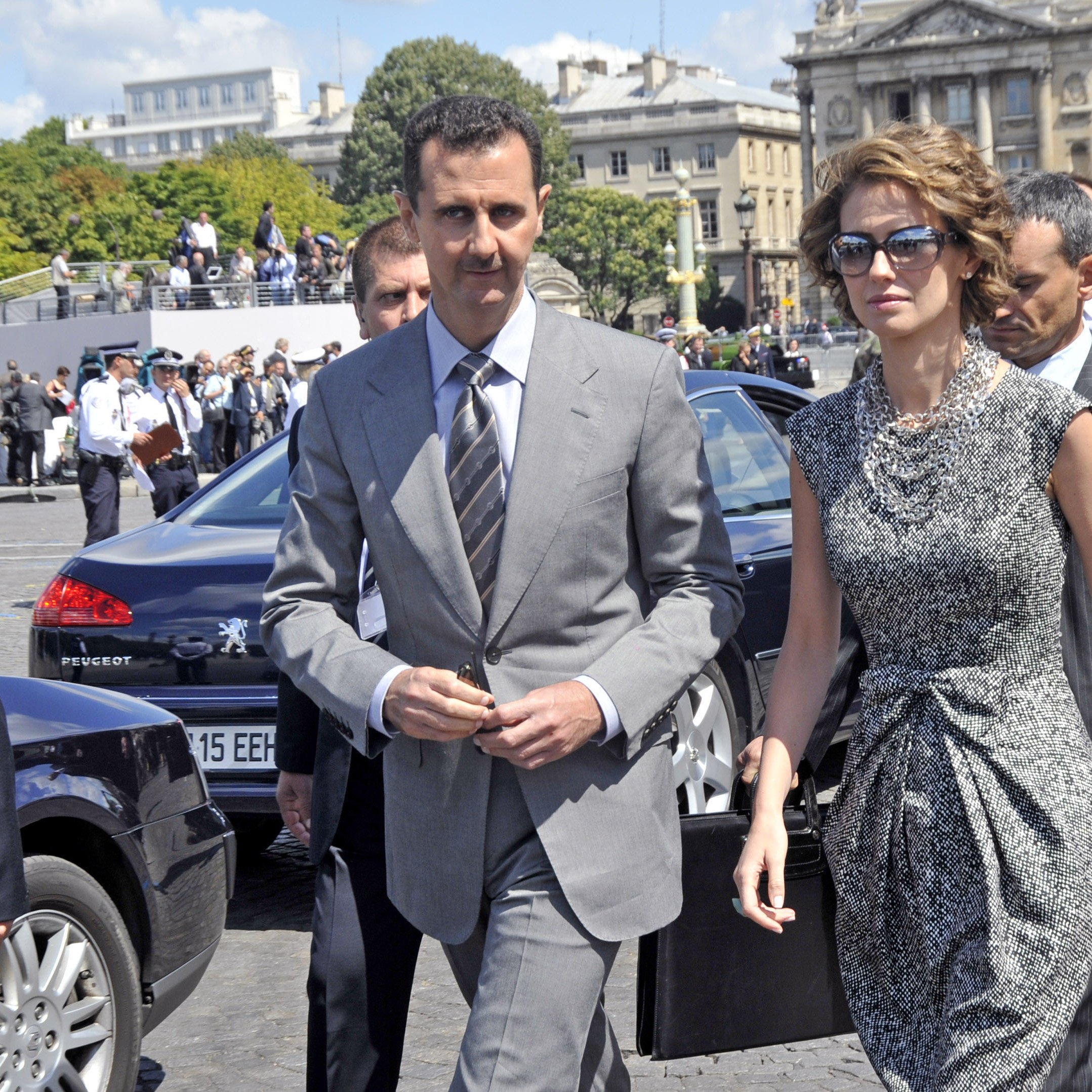 The image shows a man and a woman walking together in an outdoor setting. The man is dressed in a grey suit and tie, while the woman is wearing a stylish, patterned dress, sunglasses, and holds a handbag. They appear to be in a public area, possibly during a formal event, with a group of people in the background, including security personnel. The architecture behind them suggests a significant building or landmark.