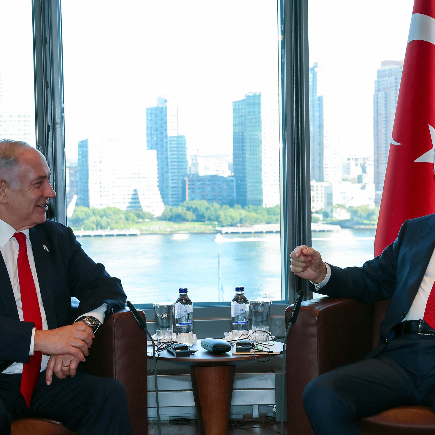 The image shows two men sitting in an office or meeting room with large windows that offer a view of a city skyline in the background. One man appears to be wearing a black suit and a red tie, while the other wears a dark suit and a white shirt with a red tie. There are symbols of Israel and Turkey nearby, indicating a diplomatic or official meeting between representatives of the two countries. The atmosphere seems cordial as they engage in conversation.