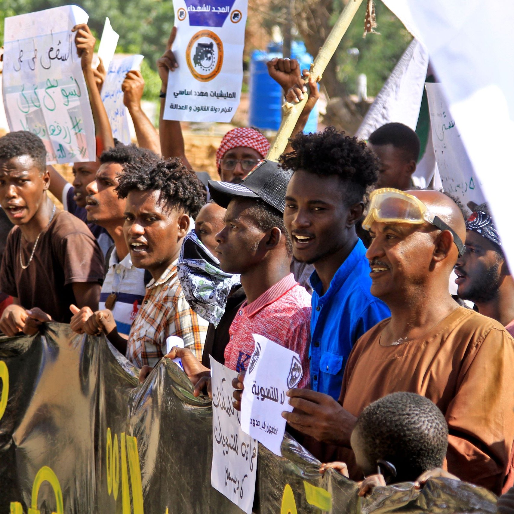 L'immagine ritrae una folla di manifestanti che tiene in mano cartelli e striscioni. I partecipanti sembrano coinvolti ed esprimono entusiasmo, alcuni sorridono mentre altri alzano le mani. Sono visibili diverse espressioni facciali e abbigliamento vario, con colori e stili che riflettono un'atmosfera di protesta. Sullo sfondo si intravede un ambiente urbano, con edifici e vegetazione. L'atteggiamento dei manifestanti suggerisce che stanno lottando per una causa importante.