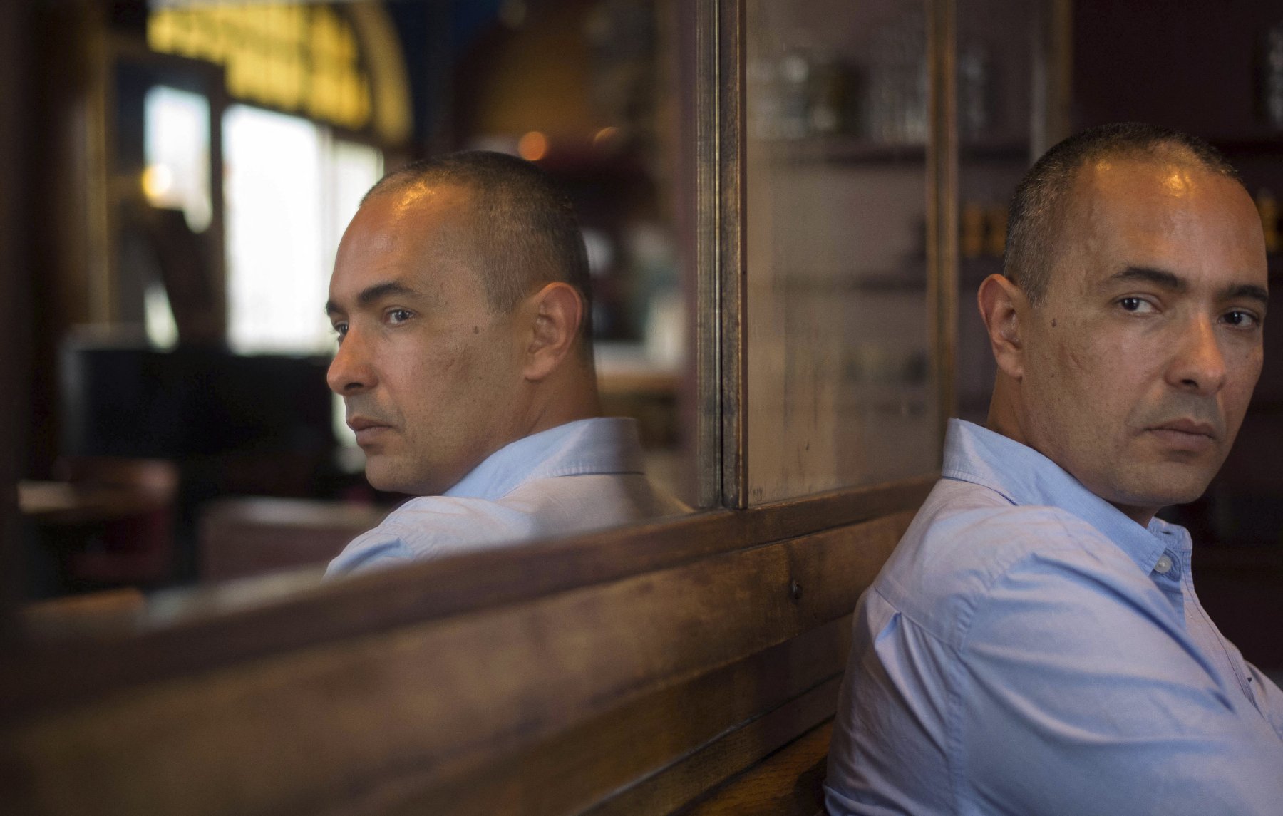 The image features a man sitting with his back to a reflective surface, creating a mirror effect. He has short, dark hair and is wearing a light blue shirt. His expression is pensive, with a serious look on his face as he gazes thoughtfully to the side. The surroundings indicate a cozy indoor setting, with warm lighting and wooden elements in the background. The reflection captures both his profile and the contemplative mood of the scene.