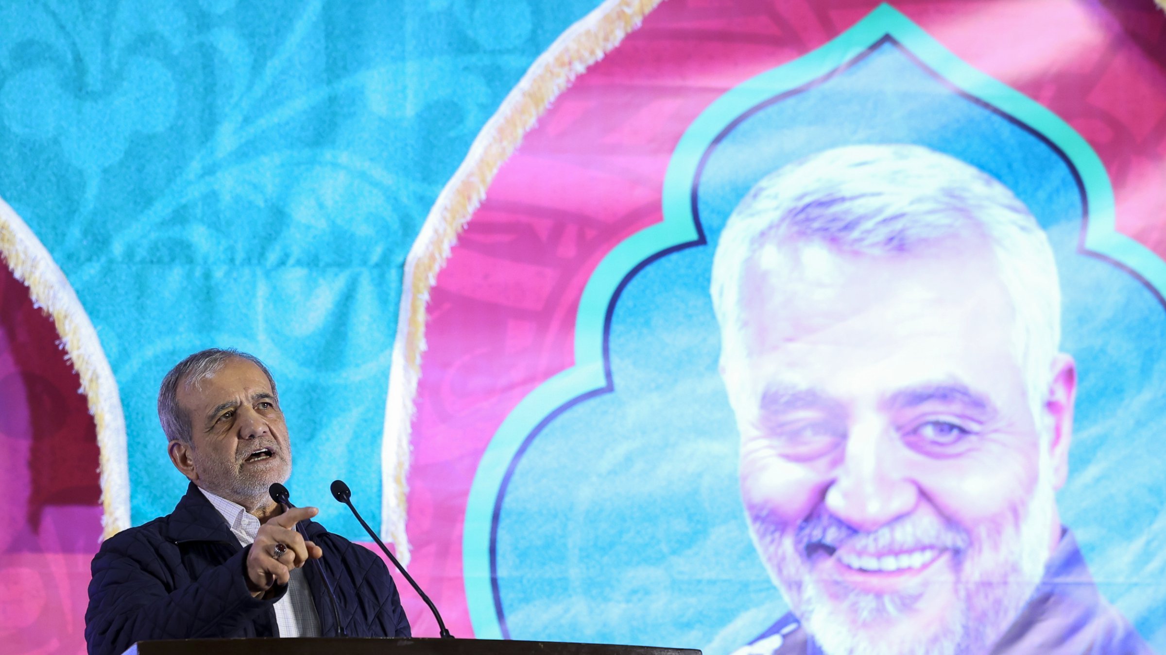 در این تصویر، شخصی در حال سخنرانی در یک مراسم دیده می‌شود. او پشت میکروفون قرار دارد و به طرف جمعیت صحبت می‌کند. در پس‌زمینه، تصویری بزرگ از فرد دیگری که لباس نظامی به تن دارد، به‌وضوح قابل مشاهده است. طراحی پس‌زمینه رنگی و با الگوهای خاص است که به مراسم زیبایی می‌بخشد. فضای کلی مراسم جدی و متعهد به هدف خاصی به نظر می‌رسد.