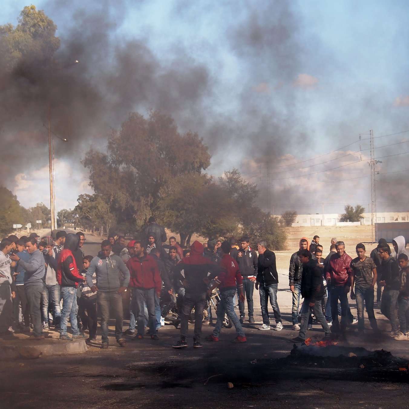 الصورة تظهر مجموعة من الأشخاص مجتمعين في منطقة مفتوحة، ويبدو أنهم في حالة من التوتر أو الاحتجاج. هناك دخان كثيف يتصاعد من حريق على جانب الطريق، مما يخلق جوًا من الاضطراب. مجموعة من الناس تجمعوا حول الحريق، ويظهر البعض منهم يرتدون ملابس مختلفة ويتجهون بنظراتهم نحو شيء ما في وسط الصورة. خلفهم يمكن رؤية أشجار وبعض المباني في المسافة، مما يشير إلى أن هذه الحادثة تحدث في منطقة حضرية أو شبه حضرية.