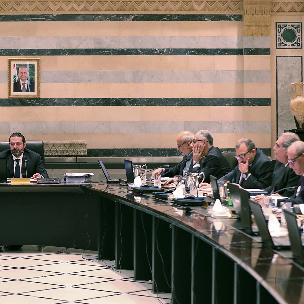 تصویر یک جلسه رسمی دولتی در لبنان را نشان می‌دهد. تعدادی از مقامات و اعضای دولت در اطراف یک میز بزرگ نشسته‌اند. در مرکز جلسه، یک مرد با لباسی رسمی و آرامش بر روی صندلی ریاست نشسته است و به دیگران نگاه می‌کند. در پس‌زمینه، پرچم لبنان و تصویری از یک شخصیت مهم بر دیوار نصب شده است. فضای اتاق با خطوط عمودی و افقی تزئینی و رنگ‌های ملایم طراحی شده است. افراد حاضر در جلسه به موضوعات مختلفی مشغول بحث و تبادل نظر هستند.