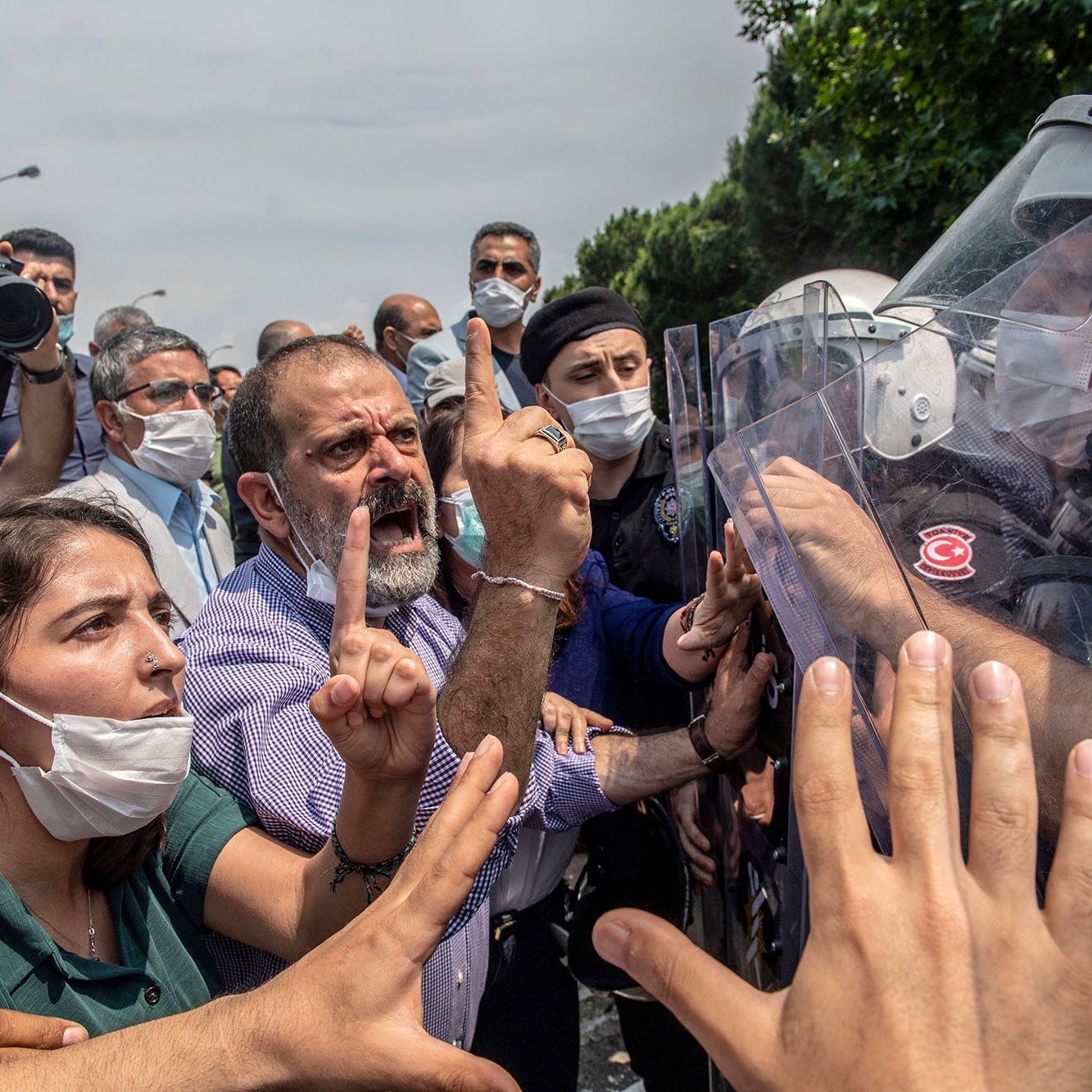 تظهر الصورة محتجين يتجمعون في مكان عام، حيث يبدو أن هناك توترًا في الأجواء. بعض الأشخاص يحملون لافتات، بينما يتواجد عدد من رجال الشرطة الذين يرتدون خوذات ودروع لمواجهة الحشد. يبدو أن هناك تفاعلًا حادًا بين المتظاهرين ورجال الأمن، مع تعبيرات وجهية تشير إلى الغضب والاحتجاج. بعض الأشخاص يرتدون أقنعة للوجه، مما يدل على احتمال وجود تدابير صحية. الأشجار والخضرة، تشير إلى أن الموقع في منطقة حضرية.