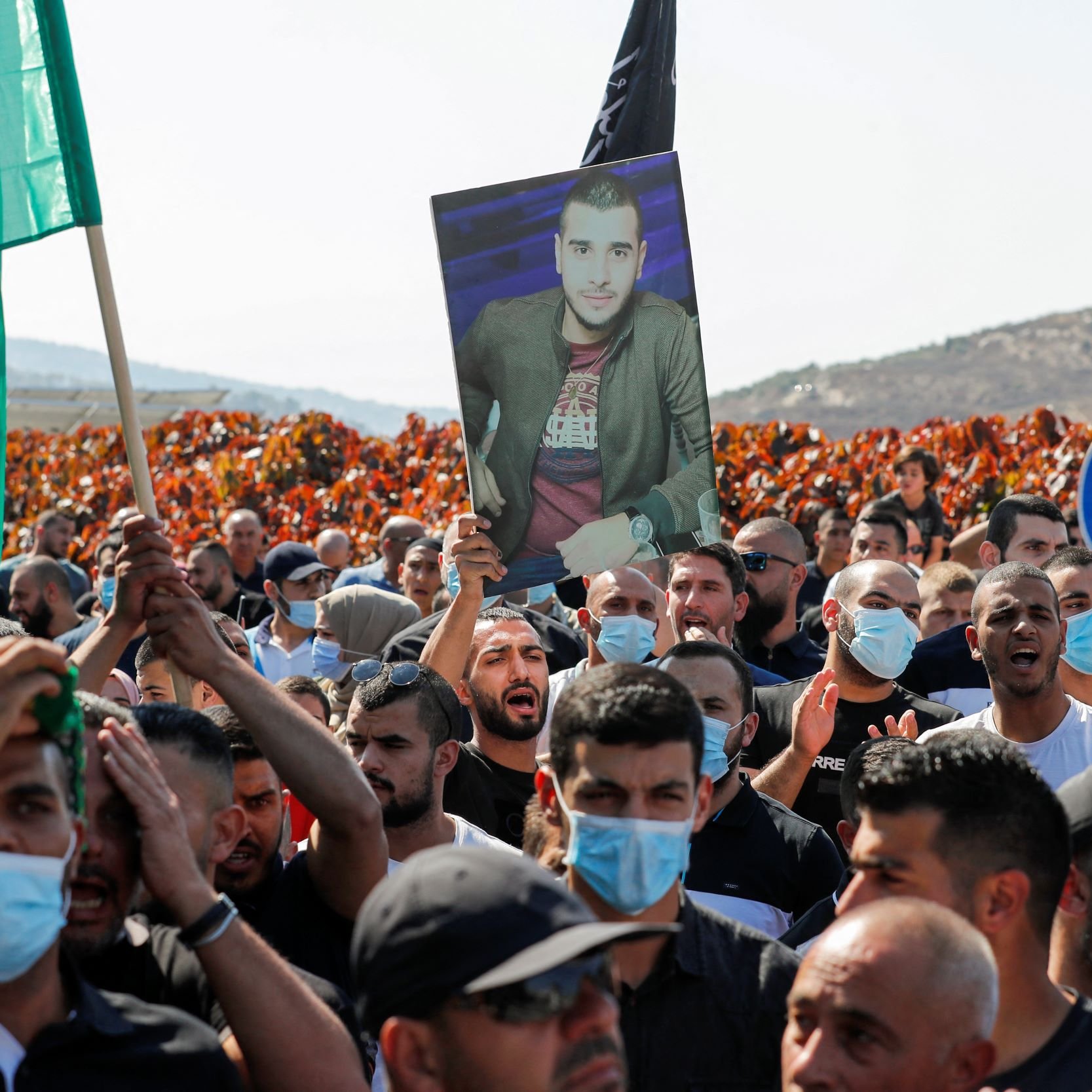 La imagen muestra a un grupo de personas en una manifestación. Hay banderas de diferentes colores ondeando, y muchos de los manifestantes llevan mascarillas. Uno de ellos sostiene una fotografía en alto de un hombre. El ambiente parece ser de protesta, y el fondo incluye áreas despejadas con vegetación. La escena transmite una fuerte sensación de unidad y determinación entre los participantes.