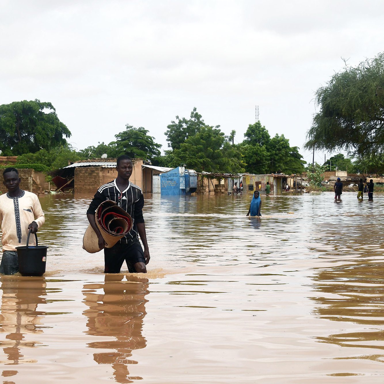 La imagen muestra una escena de inundación, donde el agua ha cubierto gran parte de un área, afectando a varias construcciones. Dos hombres caminan a través del agua, uno de ellos lleva un balde y el otro sostiene una manta enrollada. Al fondo, se pueden ver casas y árboles, todos rodeados de agua. El cielo está nublado, lo que sugiere una atmósfera sombría y húmeda. La inundación ha dejado a la comunidad en una situación difícil.