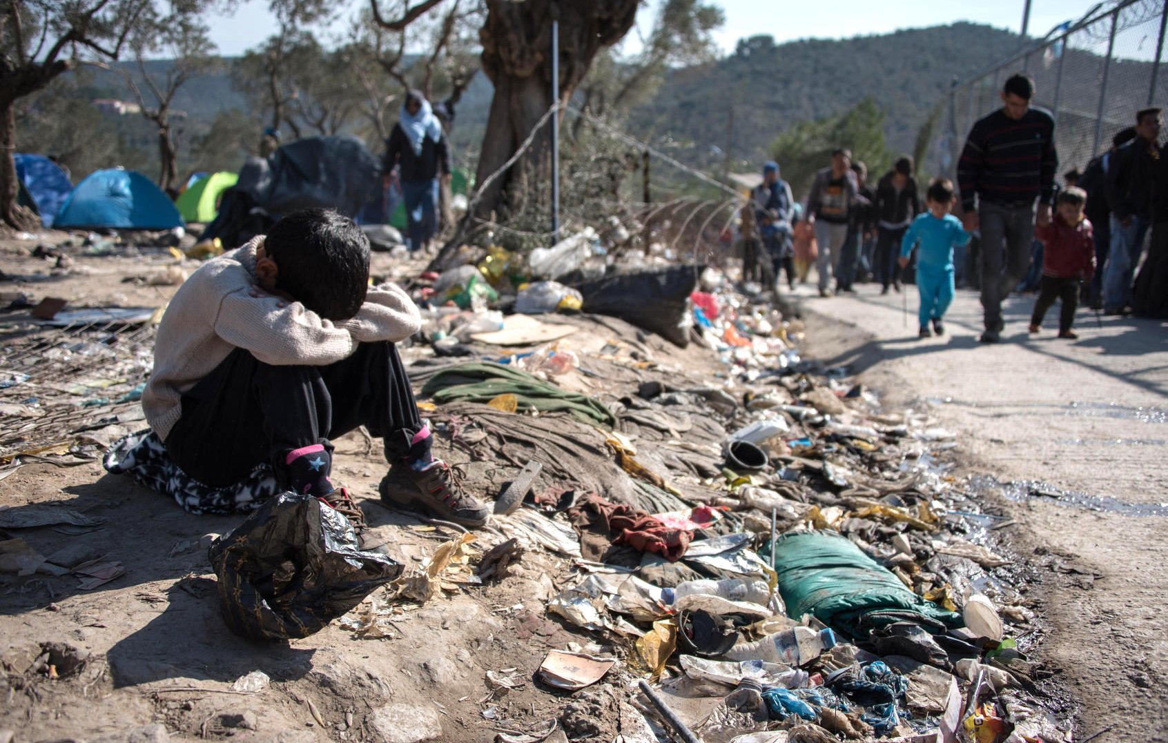 تظهر الصورة طفلاً يجلس على الأرض في ملجأ مهدم أو منطقة غير نظيفة، حيث يبدو عليه الإحباط أو الحزن، ويضع رأسه بين ركبتيه. حوله هناك أكوام من القمامة والأشياء المهملة، مما يعكس ظروف معيشة قاسية. في الخلفية، يمكن رؤية مجموعة من الأشخاص تسير في اتجاه آخر، مما يعزز الشعور بالانفصال والمعاناة. تعكس الصورة بوضوح التحديات التي يواجهها الأفراد في مثل هذه الظروف.