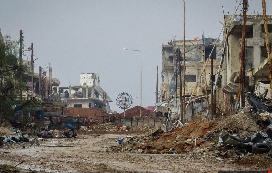 L'image montre une scène de destruction urbaine. On peut voir des bâtiments en ruines, avec des murs effondrés et des structures endommagées. Le sol est recouvert de débris, et l'atmosphère semble sombre et désolée. Il y a des poteaux électriques dans l'arrière-plan, et un éclairage public est visible au loin, mais tout cela contribue à une ambiance de désolation. La végétation est rare, et l'ensemble dégage une impression de lieu abandonné et ravagé.