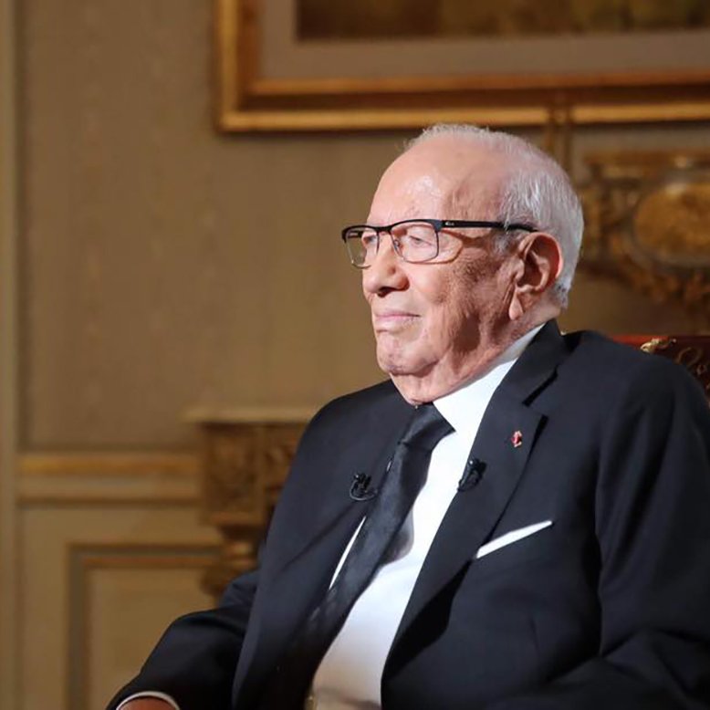 The image depicts an elderly man sitting in a formal setting. He is wearing glasses and a black suit, along with a black tie. The background features tasteful decor with intricate designs and warm colors, suggesting an elegant environment. The man appears to be engaged in thought or reflecting, with a calm and serene expression on his face.