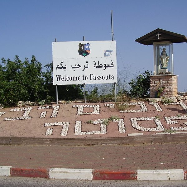 L'image montre un panneau de bienvenue à Fassouta, une localité. Le panneau est écrit en arabe et en anglais. À côté du panneau, il y a un abri qui semble abriter une statue, probablement de la Vierge Marie, indiquant une certaine importance religieuse à cet endroit. Le sol est décoré avec des mots formés de cailloux blancs, ajoutant une touche artisanale à l'ensemble. En arrière-plan, on aperçoit des arbres ou de la verdure, donnant une impression de nature environnante.