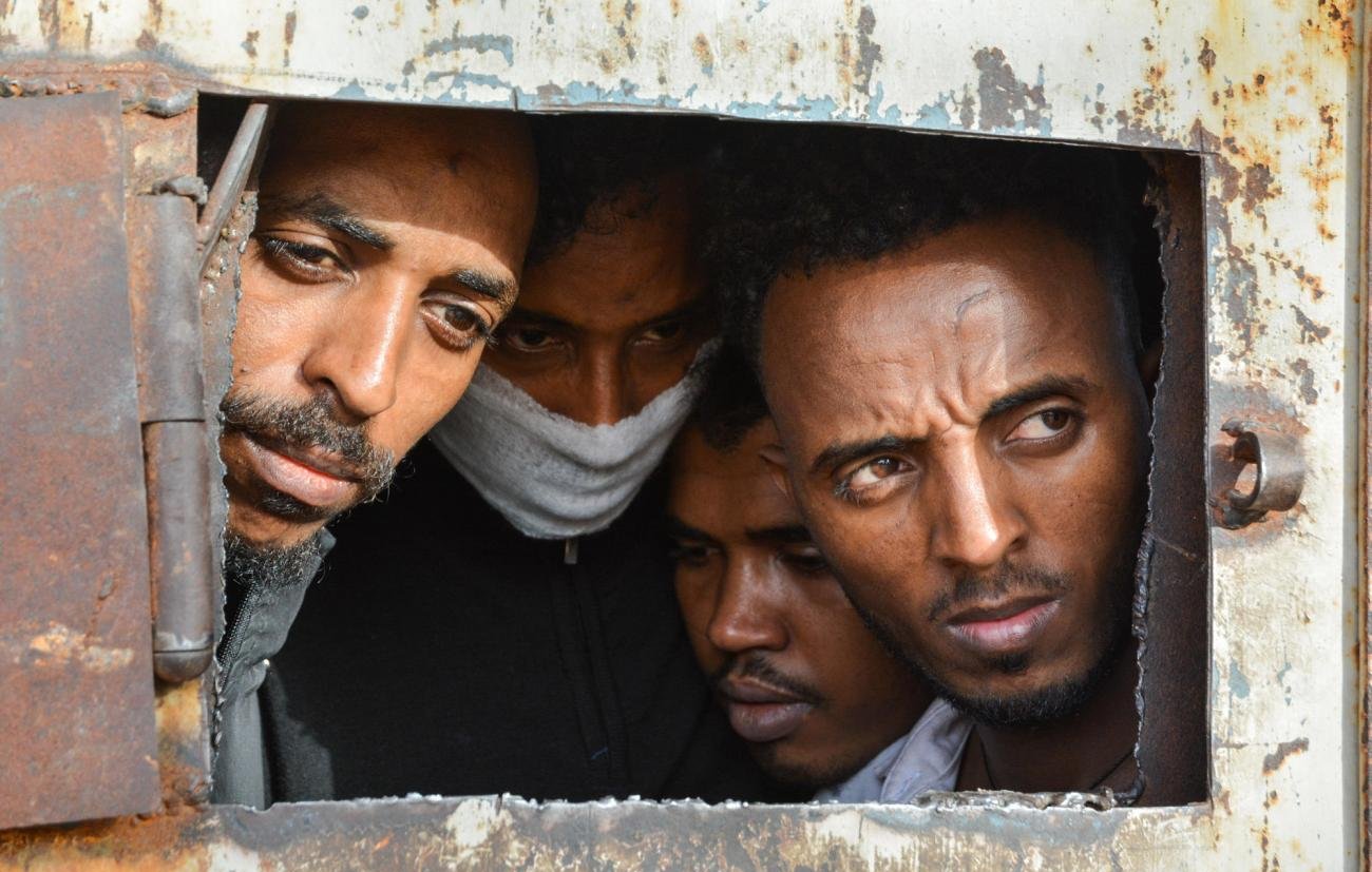 تظهر في الصورة مجموعة من الأشخاص ينظرون من خلال فتحة صغيرة في باب أو جدار صدئ. تعكس تعابير وجوههم مشاعر القلق والترقب، كما يبدو أنهم مقيدون أو محاطون بظروف صعبة. الخلفية تظهر علامات التآكل والصدأ، مما يضيف إلى الأجواء العامة للصورة.