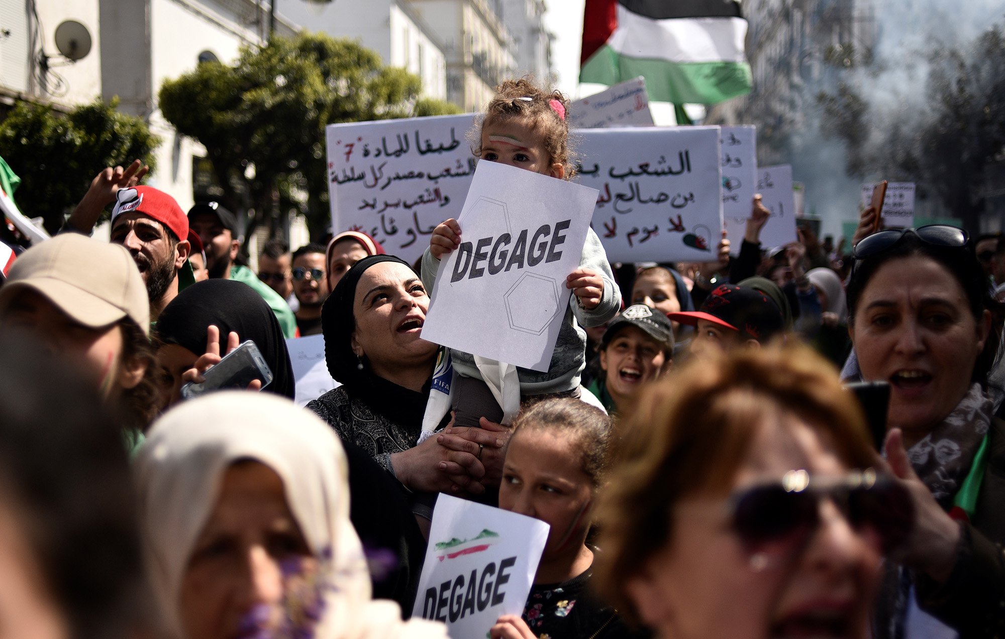 L'image montre une foule rassemblée pour une manifestation. Au premier plan, une femme porte un enfant sur ses épaules, et elle tient un panneau avec le mot "DEGAGE". Les manifestants semblent exprimer des revendications politiques. On peut également voir d'autres panneaux en arabe et des drapeaux, ce qui indique une atmosphère de protestation collective. La scène se déroule en plein air, probablement dans une ville, et les expressions des participants montrent une forte détermination.