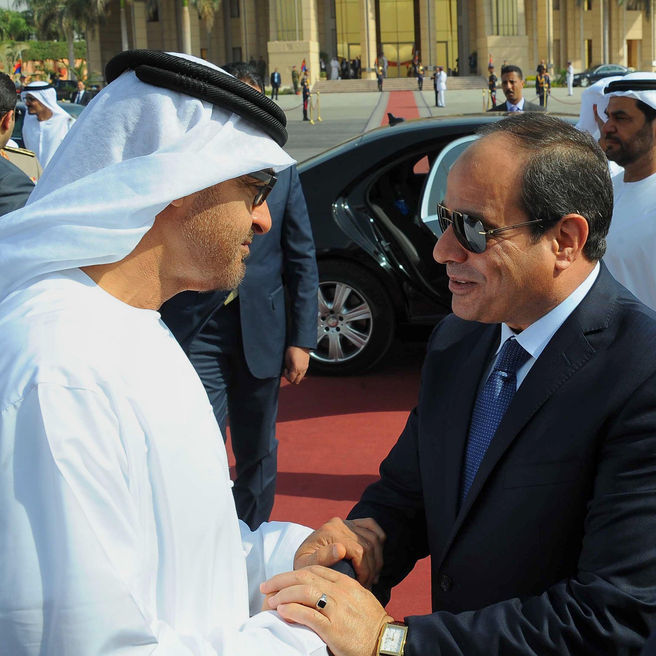 تظهر في الصورة شخصيات يرتديان ملابس تقليدية. الشخص على اليسار يرتدي الثوب الإماراتي الأبيض والغترة، بينما الشخص على اليمين يرتدي بدلة رسمية. يبدو أنهم يتصافحان في مناسبة رسمية، مع وجود سيارات فاخرة في الخلفية ومجموعة من الحراس أو الأشخاص الآخرين. تعكس الصورة أجواء من التعاون والاحترام المتبادل.