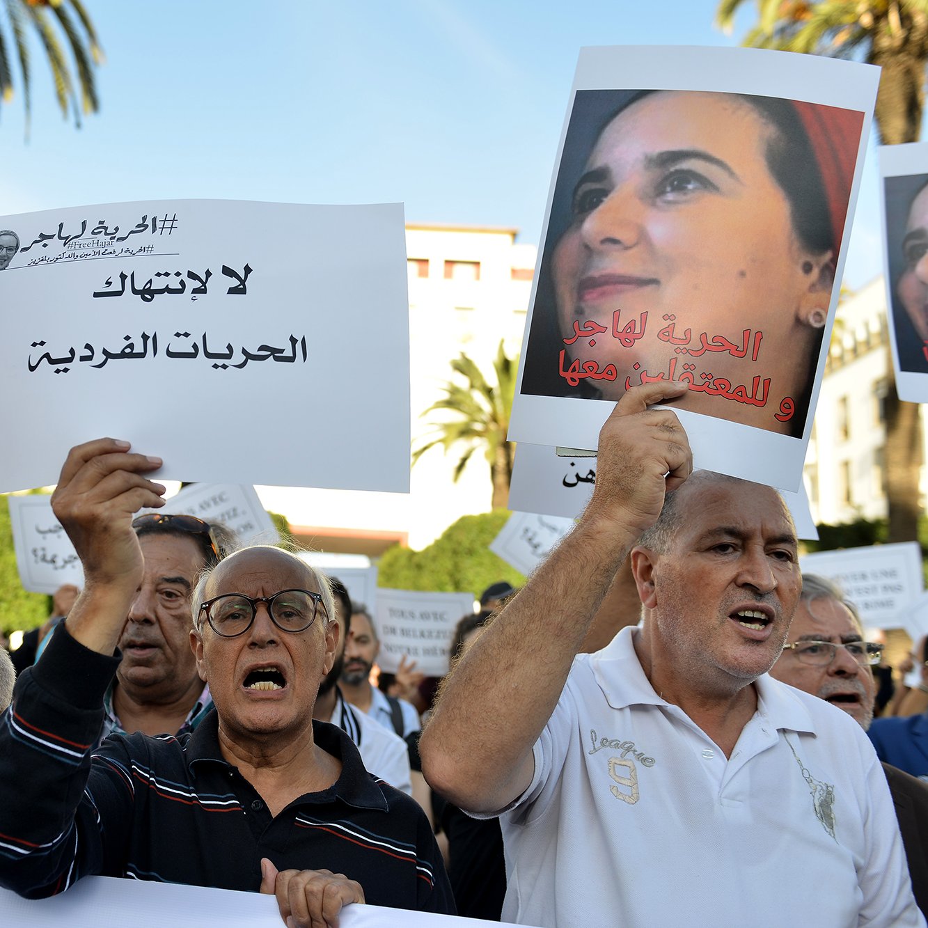 تظهر في الصورة مجموعة من الناس يتظاهرون في مكان عام، حاملين لافتات مكتوبة. يبدو أنهم يعبرون عن مطالب تتعلق بالحرية الفردية وحقوق المعتقلين. المشهد يدل على حماس المشاركين، حيث يعبر بعضهم عن استيائهم بصوت عالٍ. في الخلفية، يمكن رؤية بعض النخيل والسماء التي تحتوي على بعض السحب، مما يدل على أن المظاهرة جرت في يوم مشمس.