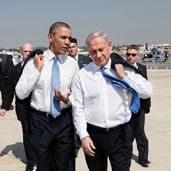 L'image montre deux hommes, habillés en costume, marchant ensemble sur un tarmac. L'un porte une cravate bleue et l'autre une cravate claire. Ils semblent engagés dans une conversation. En arrière-plan, on peut apercevoir des membres de la sécurité et des véhicules. Le ciel est clair, et l'environnement semble être un aéroport.