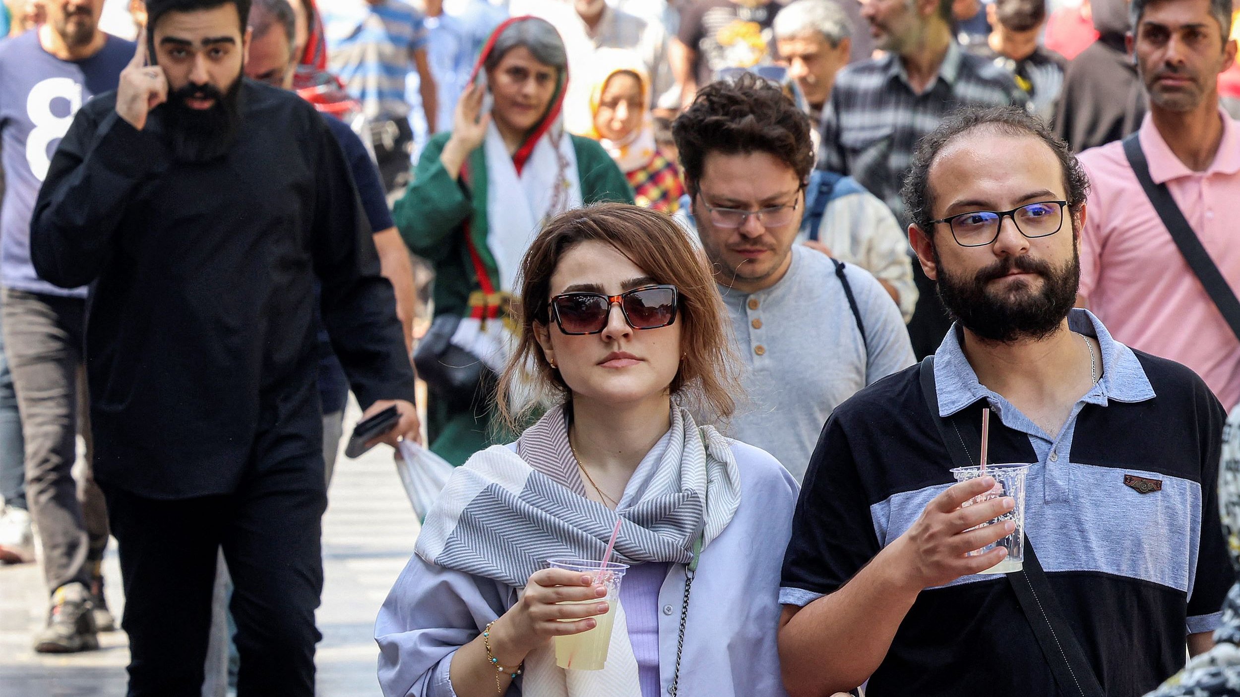 L'image montre une scène animée d'une rue où plusieurs personnes marchent. Au premier plan, deux personnes se déplacent côte à côte. Un homme portant des lunettes et une chemise à rayures tient un verre. À côté de lui, une femme avec des lunettes de soleil et une écharpe semble s'engager dans une conversation. En arrière-plan, d'autres personnes marchent, créant une atmosphère dynamique. On peut voir une variété de styles vestimentaires, suggérant une diversité culturelle.