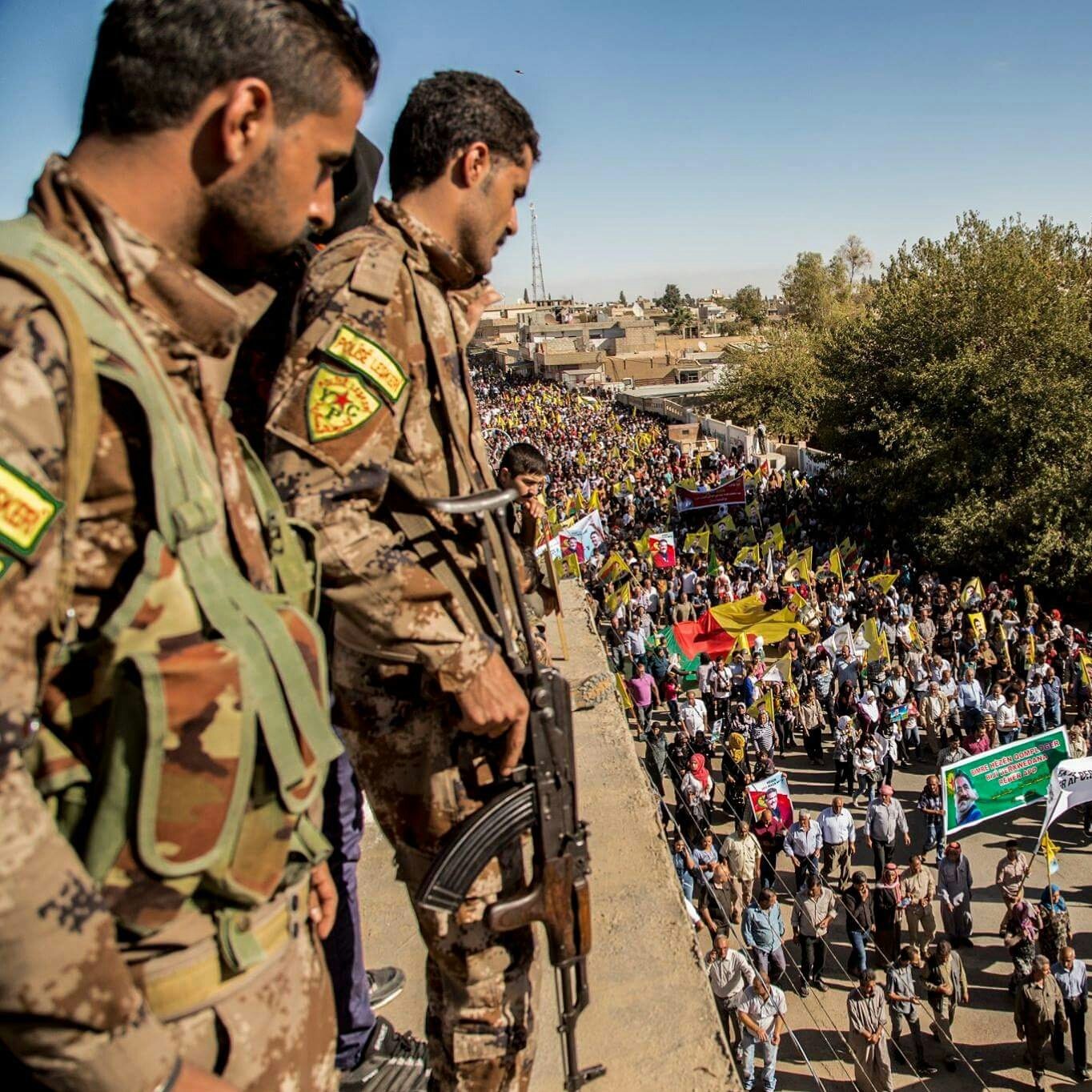 The image depicts a large crowd participating in a demonstration or march. In the foreground, two armed soldiers in military uniforms are positioned on a higher vantage point, observing the event. Below them, a diverse group of people holds various flags and banners, indicating that the gathering is likely a political or social protest. The scene is set in an outdoor urban environment, with trees and buildings visible in the background. The atmosphere appears to be one of engagement and collective action.