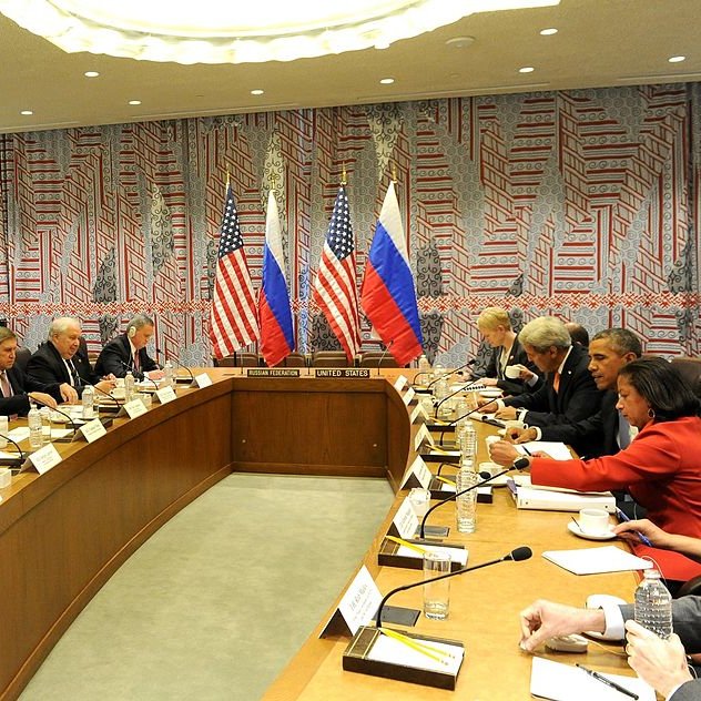 L'image montre une réunion formelle se déroulant dans une salle de conférence. À une longue table, des représentants des États-Unis et de la Russie sont présents, avec des drapeaux des deux pays visibles en arrière-plan. Les participants, qui semblent être des diplomates ou des responsables gouvernementaux, sont engagés dans une discussion sérieuse. La décoration de la salle présente des motifs géométriques sur les murs, et l'ambiance générale est celle d'une négociation ou d'un dialogue diplomatique.