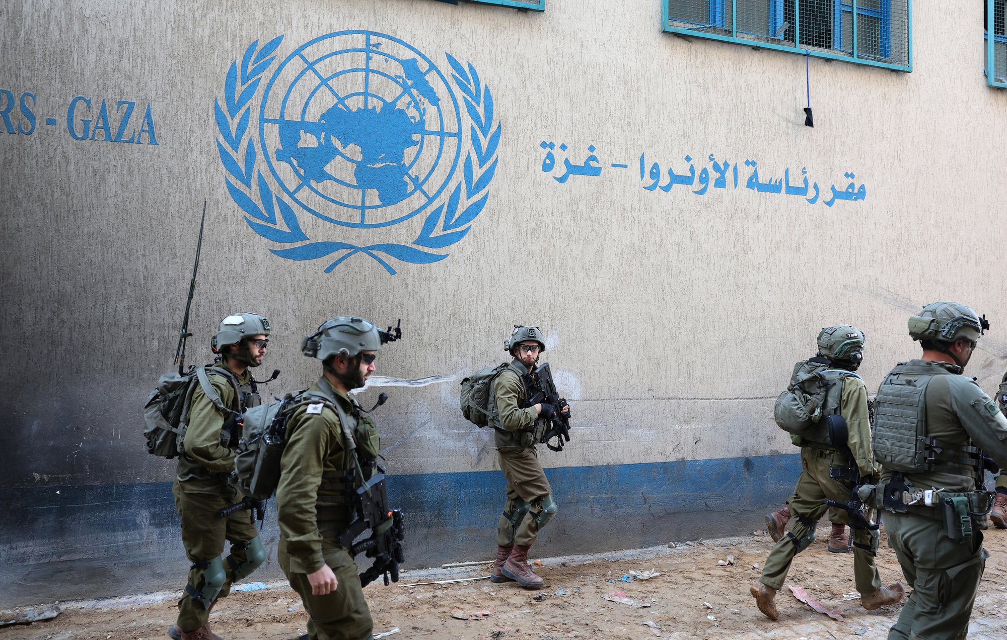 L'image montre des soldats en uniforme marchant près d'un bâtiment, probablement associé à l'ONU, avec un logo visible sur le mur. Leurs tenues militaires indiquent qu'ils sont en mission. L'environnement semble urbain, avec des structures typiques d'une zone habitée. L'atmosphère peut être perçue comme tendue en raison de la présence militaire.