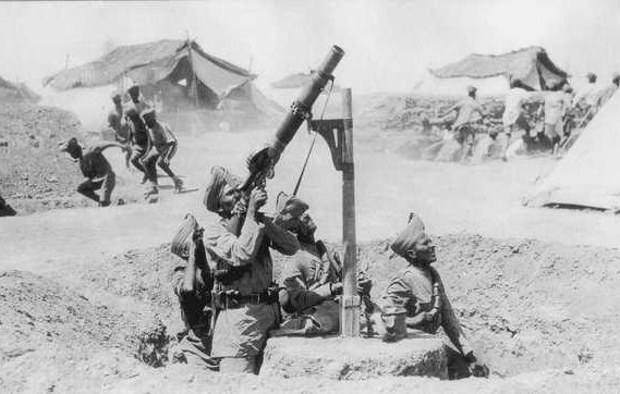 The image depicts a group of soldiers operating a mortar in a wartime setting. They are wearing military uniforms and are positioned in a trench-like area. One soldier is aiming the mortar, while other soldiers are present nearby, possibly providing support or preparing for an attack. In the background, there are tents suggesting a military camp, and additional soldiers can be seen, likely engaged in activity related to the ongoing conflict. The photo has a historical, black-and-white appearance, conveying a sense of the era in which it was taken.