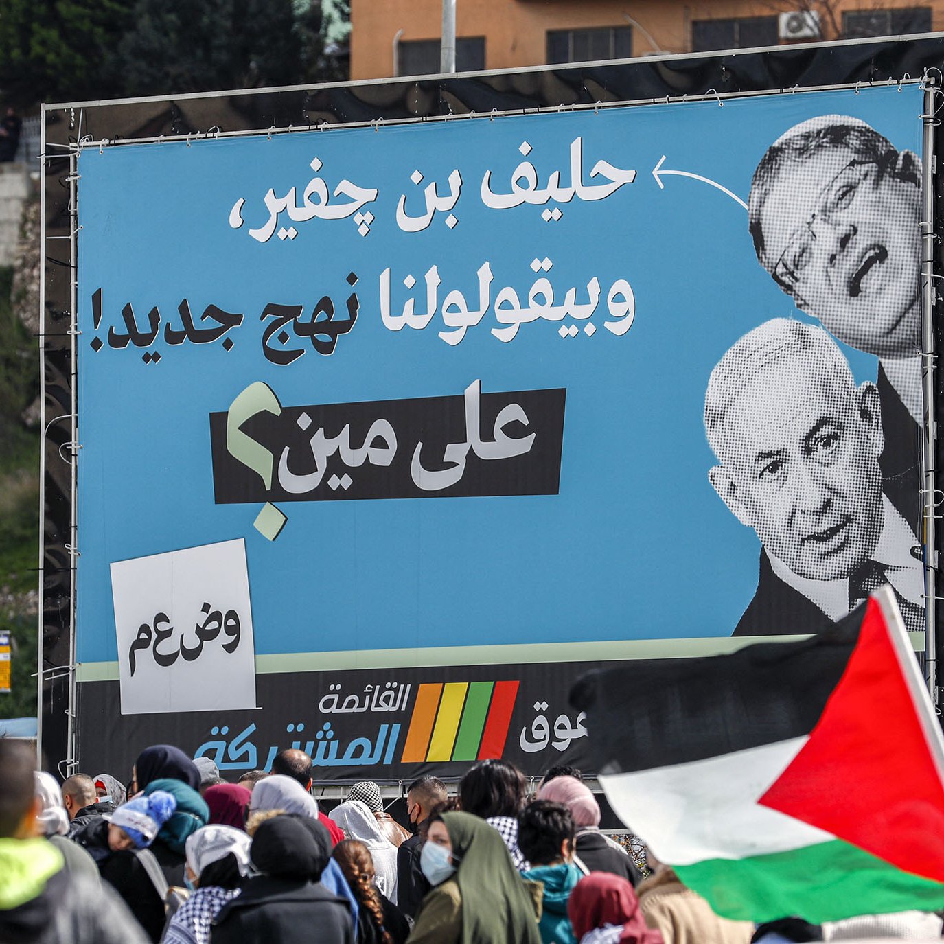 La imagen muestra una gran pancarta en un evento, donde destacan las imágenes de dos personas, una de ellas es un hombre que parece ser una figura política conocida. El cartel contiene texto en árabe, con un mensaje crítico, probablemente relacionado con la situación política. En primer plano, se observan personas reunidas, algunas de las cuales sostienen banderas, sugiriendo que puede ser una manifestación o protesta. La atmósfera parece ser de movilización social.