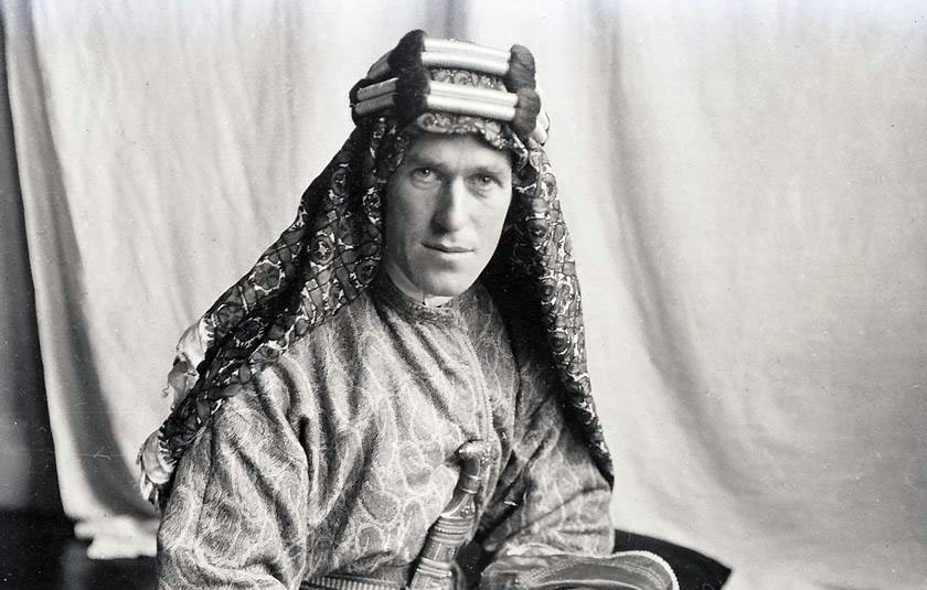 L'image montre un homme portant un costume traditionnel. Il est vêtu d'une tunique ornée et d'un headdress décoratif. Son regard est sérieux et il tient un livre dans ses mains. L'arrière-plan est sobre, ce qui met en valeur son apparence et son habillement. La photo semble être ancienne, ce qui lui donne un aspect historique.