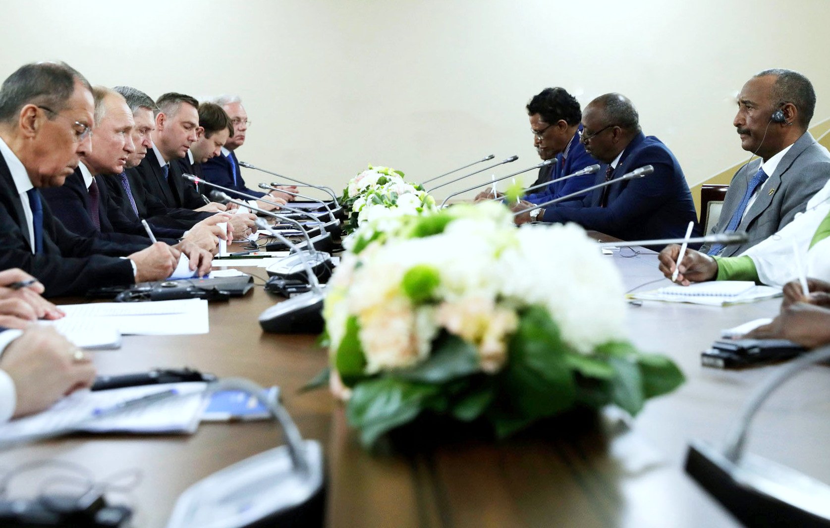 The image depicts a formal meeting or conference, with a long table where several individuals are seated across from each other. There appears to be a floral centerpiece on the table. The participants seem to be engaged in discussion or note-taking, with some looking attentively at documents or each other. The setting appears to be a neutral, professional environment, likely a government or diplomatic meeting.