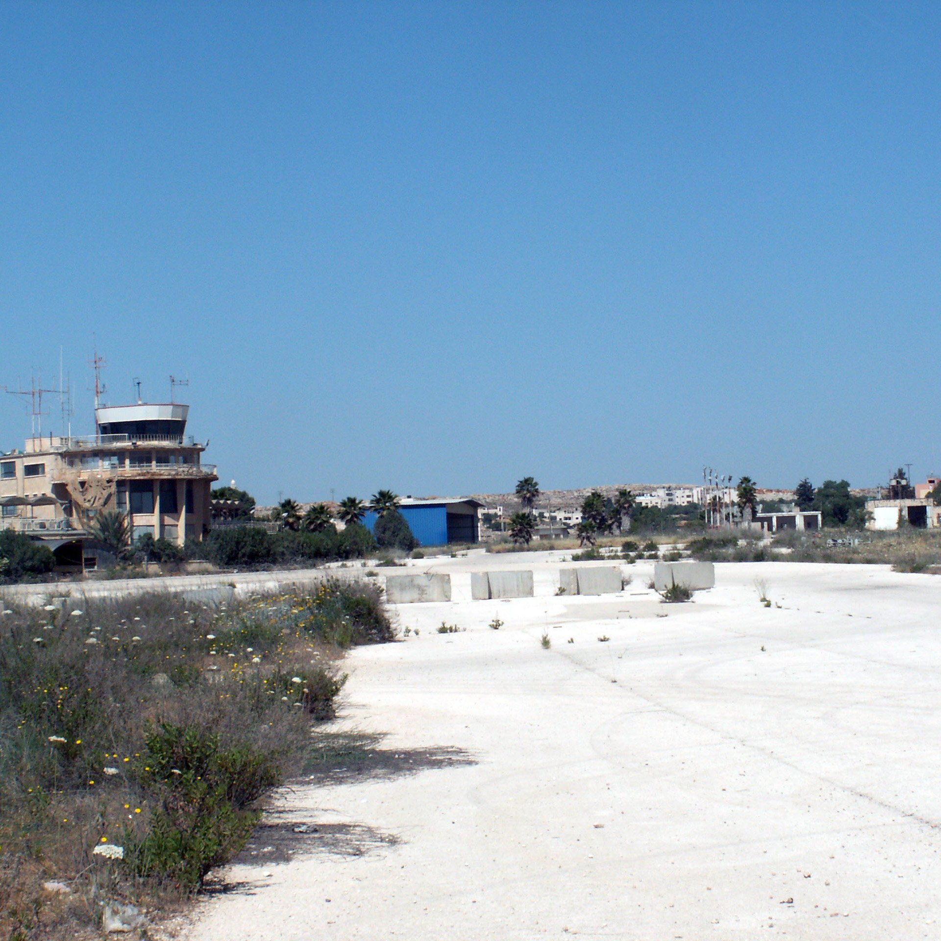 L'image montre un paysage de terrain vague, probablement une ancienne installation ou un aéroport désaffecté. Au loin, on peut voir un bâtiment de type tour de contrôle, avec des antennes sur le toit. Le sol est principalement fait de béton, avec des herbes et des fleurs sauvages qui poussent entre les fissures. Le ciel est dégagé et d'un bleu intense, et en arrière-plan, d'autres structures peuvent être aperçues, suggérant un site qui n'est plus en activité. L'atmosphère semble calme et quelque peu abandonnée.