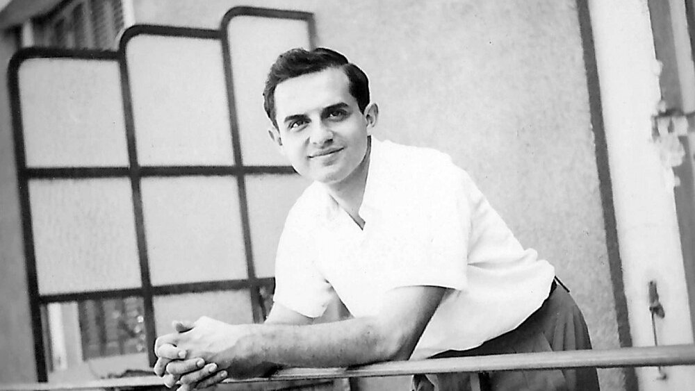 الصورة تظهر شابًا واقفًا على شرفة، يبدو مبتسمًا ومسترخيًا. يرتدي قميصًا أبيض وبنطالًا طويلًا. الخلفية تتضمن حائطًا وبعض التفاصيل المعمارية مثل النوافذ والحواجز. تعكس الصورة أجواءً من السعادة والبساطة.