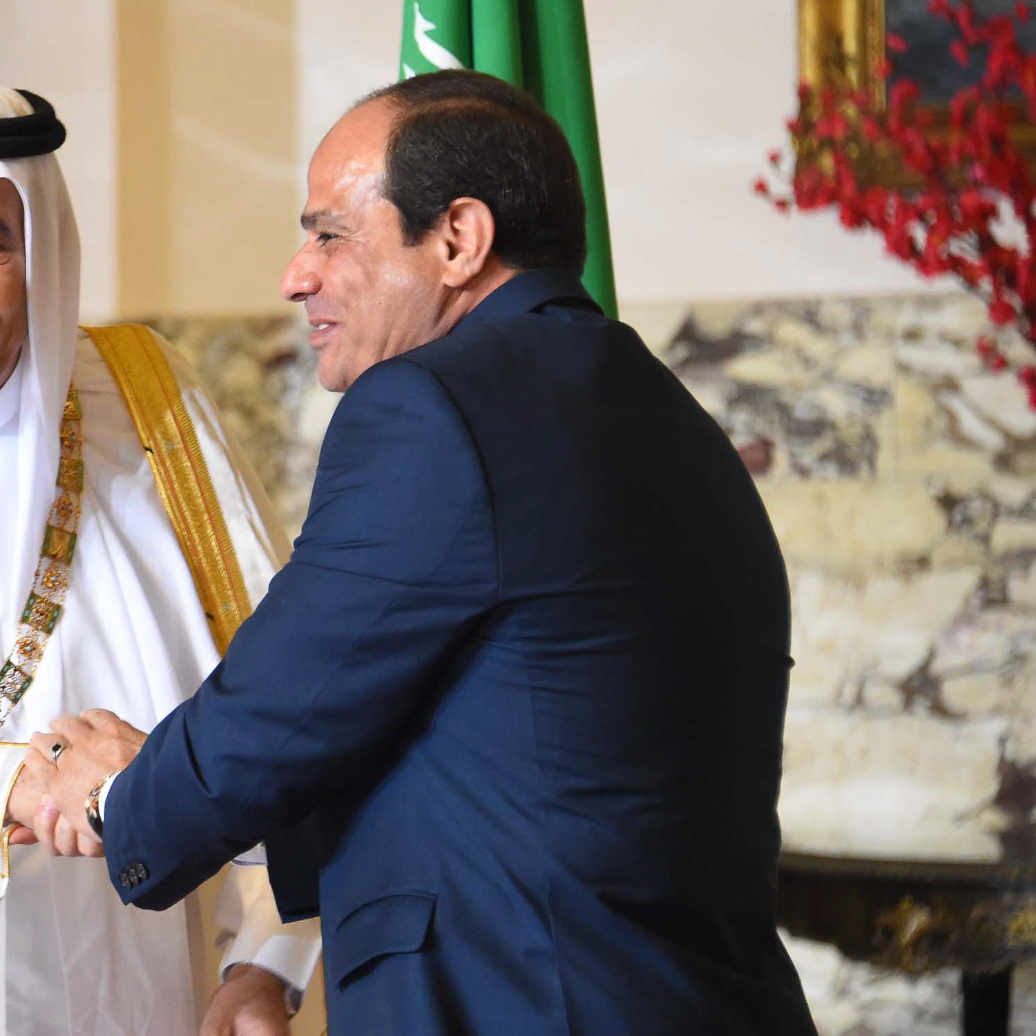 The image depicts two men in formal attire shaking hands. One man is wearing traditional Saudi clothing, including a white thobe and a black and white headscarf, adorned with a gold sash. The second man is dressed in a dark suit. They appear to be engaged in a cordial greeting, likely in a diplomatic or official context. In the background, there are hints of decor that suggest an elegant setting, possibly within a government building. One of the men is also positioned near a flag, indicating a connection to national representation.