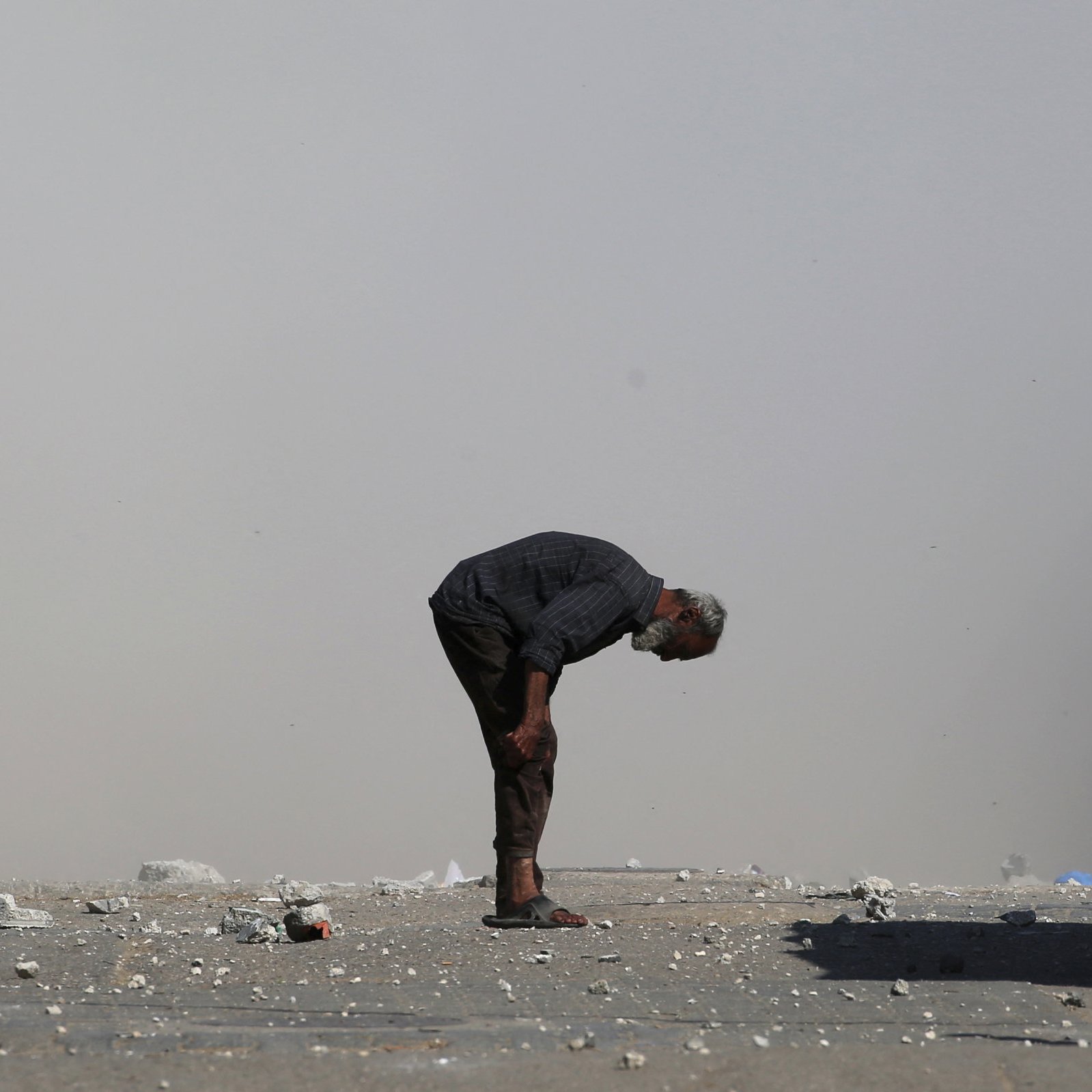 Un uomo anziano si piega in avanti su una strada polverosa, circondato da detriti.