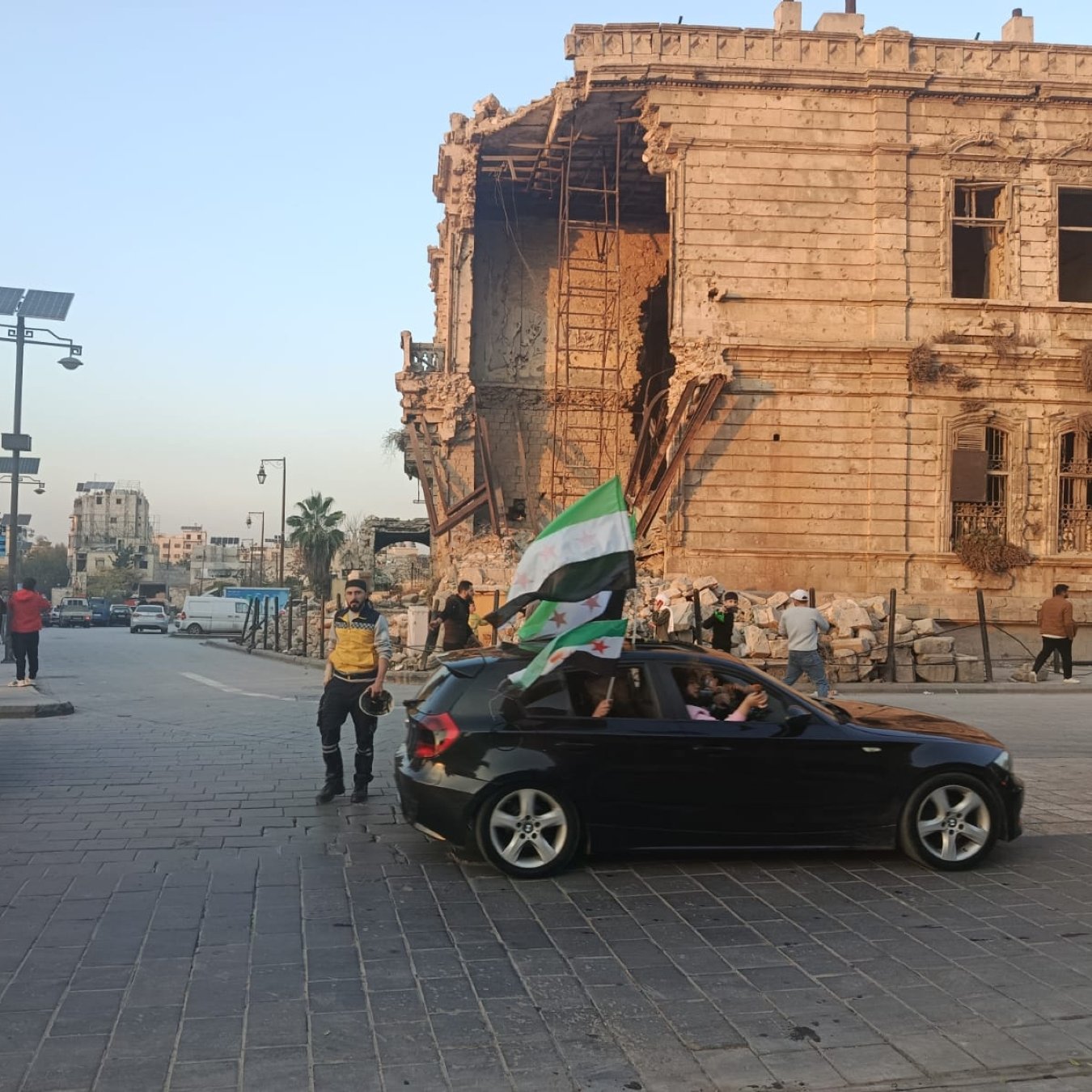 Scène de rue avec un bâtiment endommagé, des gens et une voiture avec un drapeau.