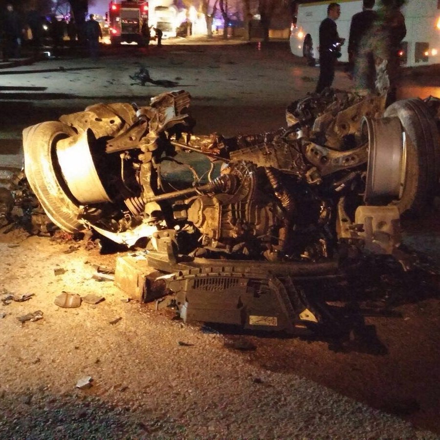 The image depicts a severely damaged vehicle that has been overturned. The car's chassis and engine are exposed, showing significant destruction, with debris scattered around the area. The scene appears to be dark, possibly at night, with some emergency responders or bystanders in the background, indicating that it may have been the site of an accident. The overall context suggests a serious incident involving the vehicle.