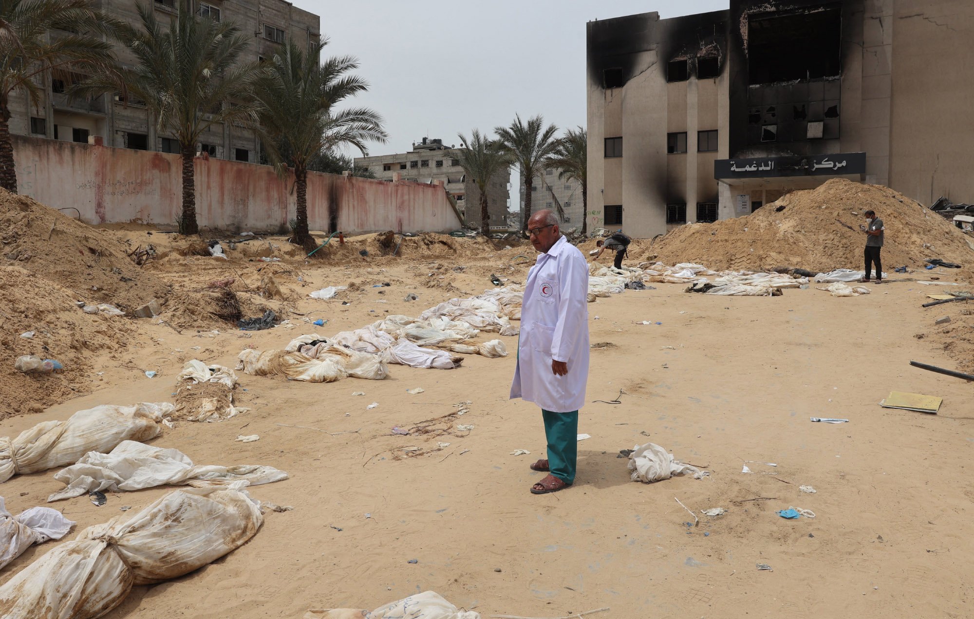L'immagine mostra un paesaggio desolato e drammatico, caratterizzato da un terreno sabbioso. Al centro, un uomo in un camice bianco osserva un'area circondata da edifici danneggiati, con alcuni segni di distruzione visibili sullo sfondo. Intorno a lui ci sono sacchi bianchi disposti a terra, che suggeriscono una scena di grande sofferenza e perdita. Palme si ergono sullo sfondo, ma l'atmosfera generale è segnata da un senso di tristezza e desolazione.