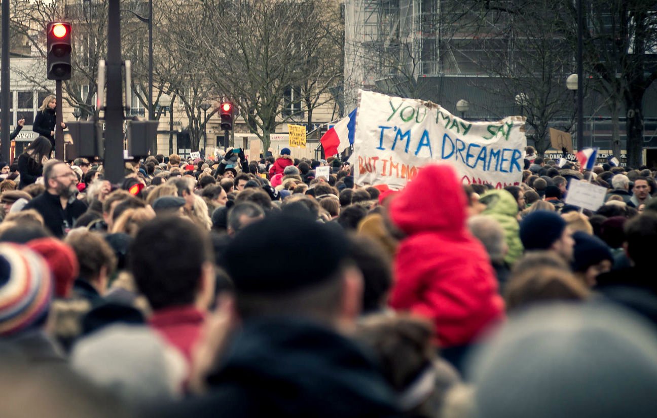 L'image montre une grande foule rassemblée, probablement pour une manifestation ou une marche. On peut voir des personnes de tous âges, certaines portant des écharpes ou des bonnets, tandis que d'autres sont vêtues de manteaux. Au centre, il y a une pancarte sur laquelle est écrit : "YOU MAY SAY I'M A DREAMER BUT I'M NOT THE ONLY ONE". Des drapeaux français sont visibles, ce qui suggère un contexte patriotique ou une revendication sociale. L'atmosphère semble être à la fois solide et engagée.