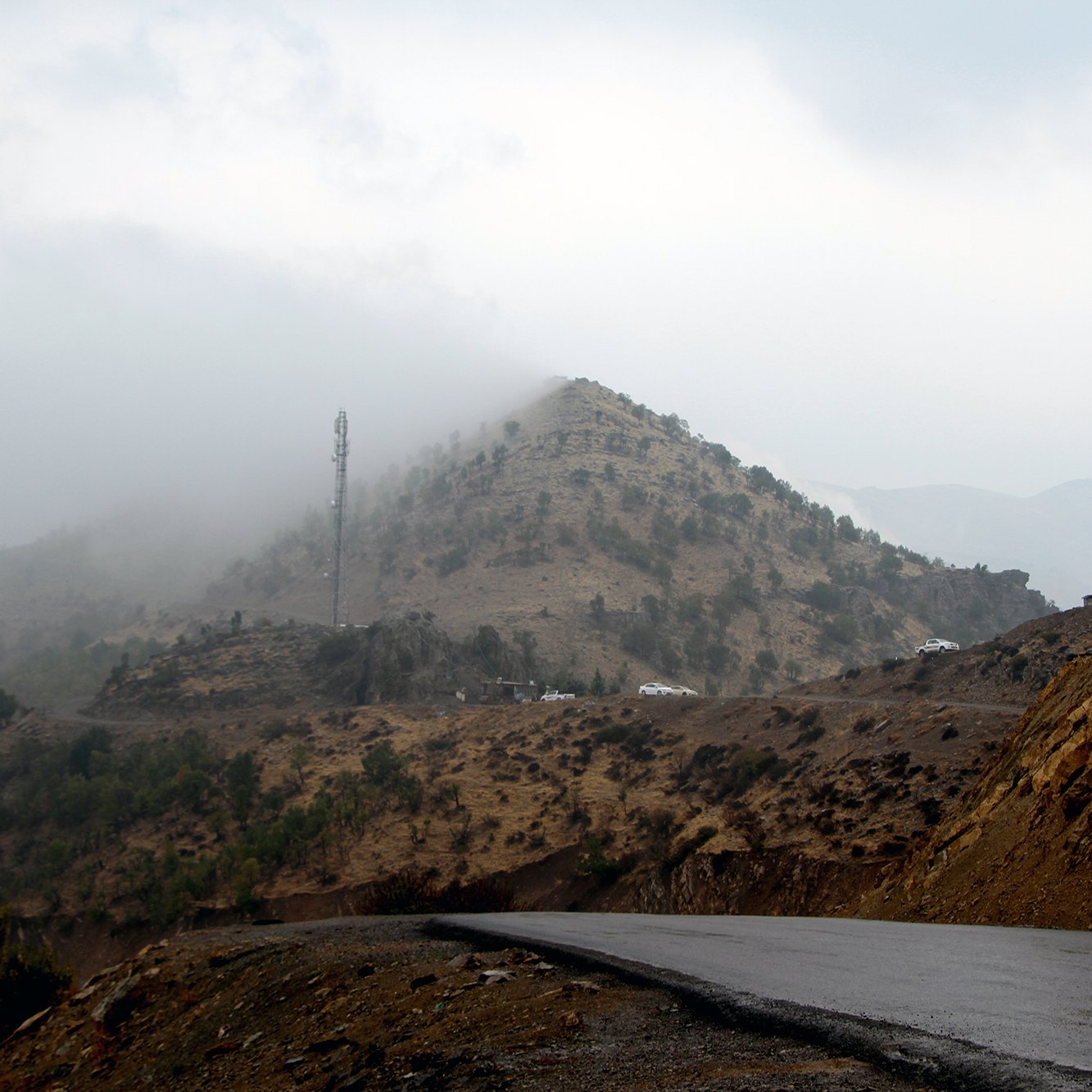 La imagen muestra un paisaje montañoso con una carretera curvada en primer plano. Al fondo, se puede ver una colina cubierta de vegetación escasa y neblina, que le da un aspecto misterioso. En la parte superior de la colina, hay una torre de comunicación. El clima parece nublado y húmedo, creando un ambiente atmosférico y sombrío.