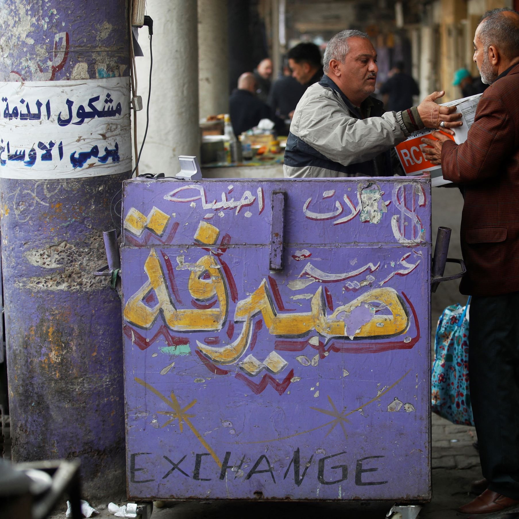 در تصویر، یک مرد در حال تبادل اطلاعات یا کالا با شخص دیگری است. دو مرد در جلوی یک دکه که بر روی آن نوشته‌هایی به زبان عربی و انگلیسی وجود دارد، ایستاده‌اند. دکه به نظر می‌آید که مربوط به تبادل کالا یا خدمات است. محیط اطراف شلوغ و بازاری به نظر می‌رسد که نشان‌دهنده زندگی روزمره در یک محل شهری است. رنگ‌های دکه روشن و نوشته‌ها واضح هستند.