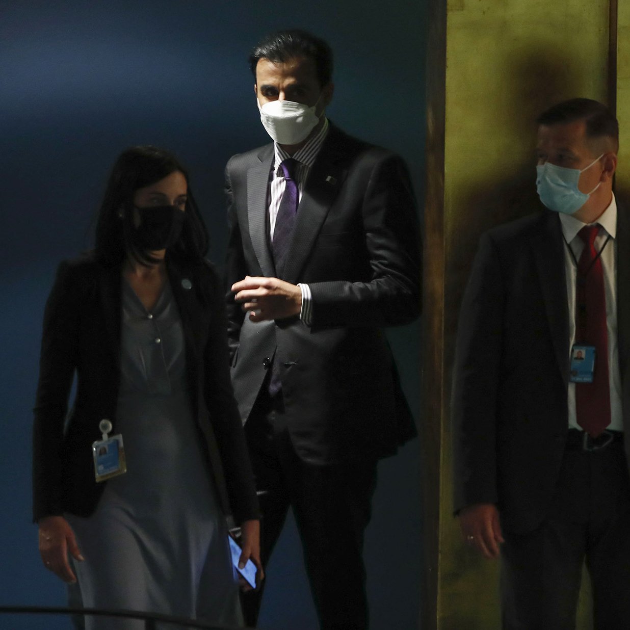 La imagen muestra a tres personas en un entorno que parece ser un edificio oficial, probablemente en una reunión o evento. Dos de ellos llevan mascarillas y lucen formales, acomodándose a un ambiente institucional. Una mujer está a la izquierda, con un vestido gris y una identificación, mientras que un hombre en el centro, vestido de traje, parece estar conversando o aguardando. A la derecha, hay otro hombre también en traje, observando. El fondo tiene colores oscuros y brillantes, sugiriendo una atmósfera seria y profesional.