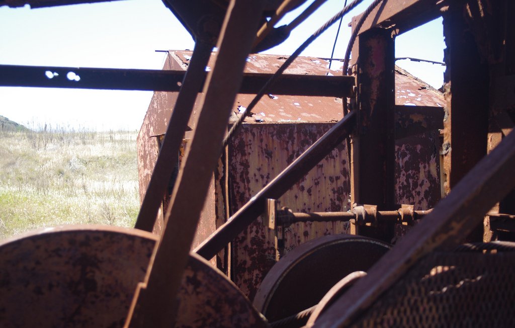 L'image montre une vue d'une structure métallique, probablement une machine ou un équipement abandonné, avec des surfaces rouillées et en décomposition. On peut voir des éléments mécaniques, tels que des poulies ou des engrenages, qui sont partiellement visibles à travers des parties de la machine. En arrière-plan, on distingue un paysage de prairies sèches, ce qui évoque une ambiance d'abandon et de dégradation. La lumière naturelle met en valeur les textures rouillées du métal.