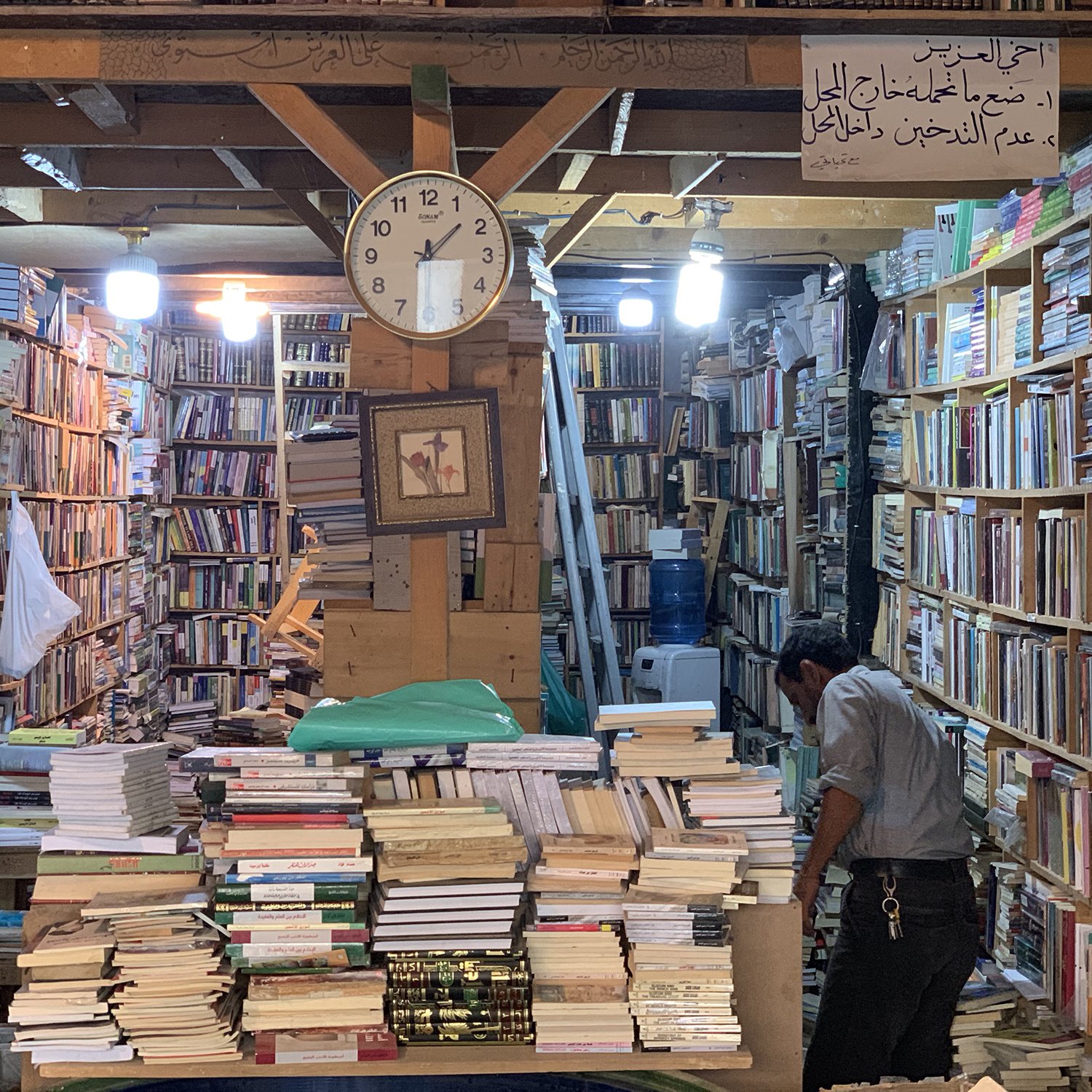 تظهر الصورة مشهداً داخلياً لمكتبة قديمة. تتراص الكتب على الرفوف الخشبية بشكل كثيف، مع ظهور بعض الكتب المكدسة على الأرض. في منتصف الصورة، يوجد ساعة حائط تشير إلى الوقت. كما يوجد شخص في الجهة اليمنى من الصورة، يبدو أنه يتفحص الكتب. الأضواء تتدلى من السقف، مما يضفي جواً دافئاً على المكان. في الأعلى، توجد لافتة مكتوبة توضح معاني تتعلق بالمكتبة.