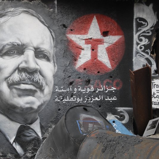 L'image montre un graffiti sur un mur, représentant un portrait en noir et blanc d'un homme avec une moustache et un regard déterminé. À côté du portrait, on peut voir une étoile rouge avec un symbole. Des inscriptions en arabe sont également visibles, accompagnées de motifs colorés sur le mur. L'ensemble donne une impression d'engagement politique, avec une ambiance urbaine et artistique.