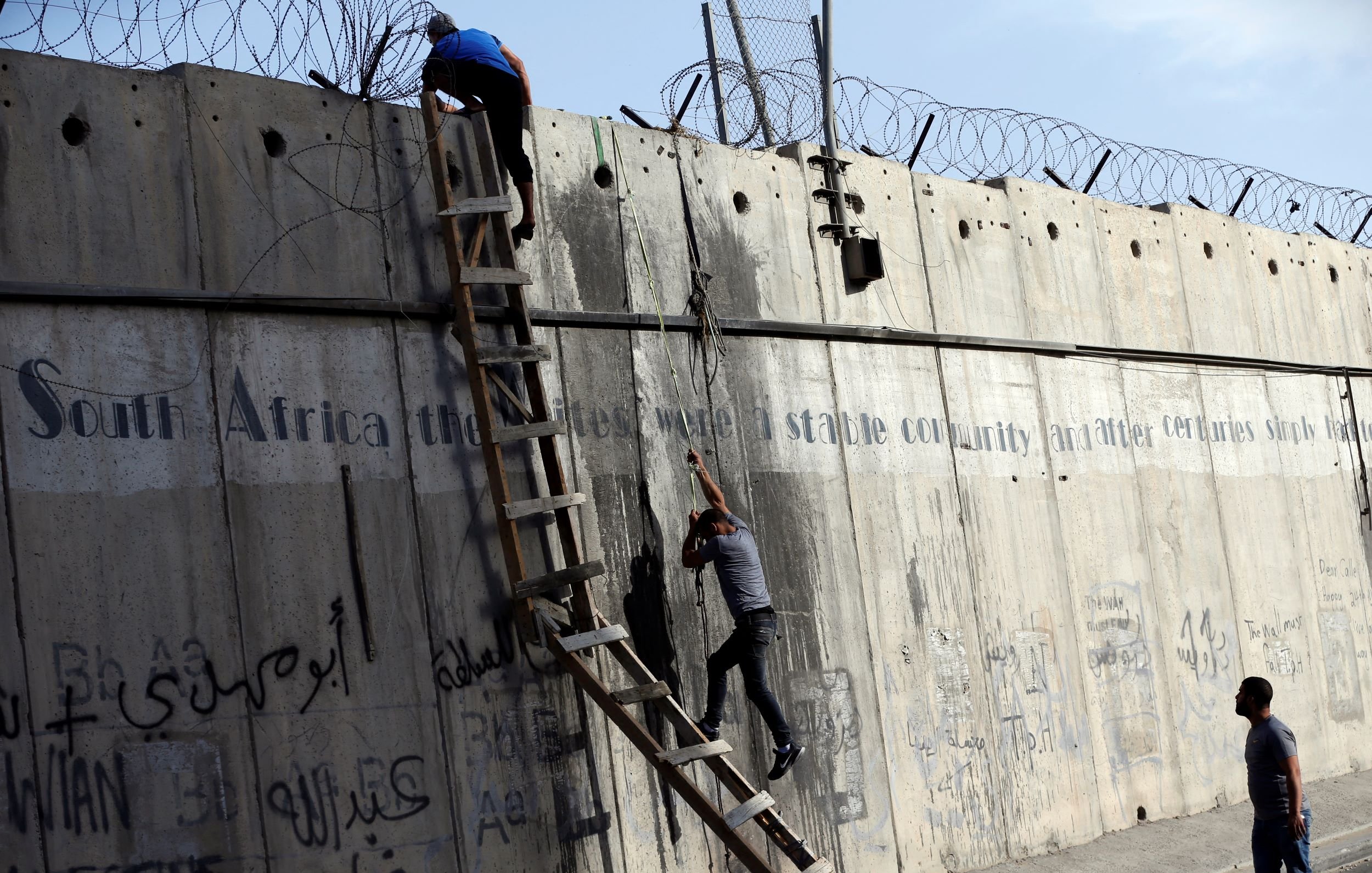 The image shows two individuals climbing a tall concrete wall, which features barbed wire at the top. A ladder is positioned against the wall, aiding their ascent. The wall bears graffiti, including text in multiple languages, and there is a phrase that mentions "South Africa" likely in the context of stability and historical struggles. The setting appears tense, capturing a moment of movement against a backdrop of urban infrastructure.