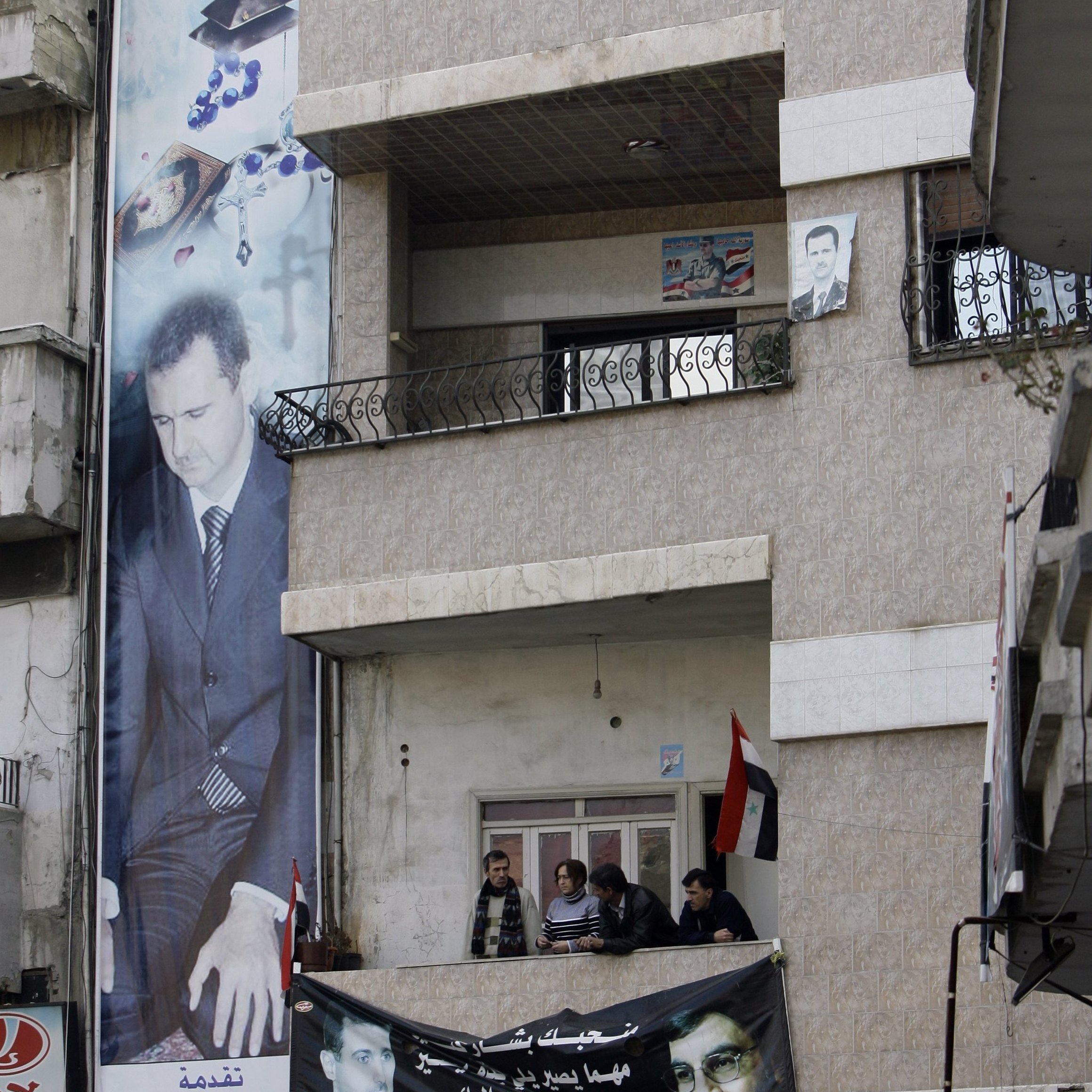 در این تصویر، نمایی از یک ساختمان چند طبقه دیده می‌شود که در آن پوسترها و تصاویر بزرگی از یک شخص (که احتمالاً یک شخصیت سیاسی است) نصب شده است. دو مرد در بالکن طبقه دوم ایستاده‌اند و به فضای بیرون نگاه می‌کنند. همچنین، در زیر ساختمان، بنرهایی با شعارها و تصاویری از دیگر افراد نصب شده است. محیط به نظر می‌رسد که در یک منطقه شهری باشد و برخی نشانه‌های ملی مانند پرچم در تصویر دیده می‌شود.